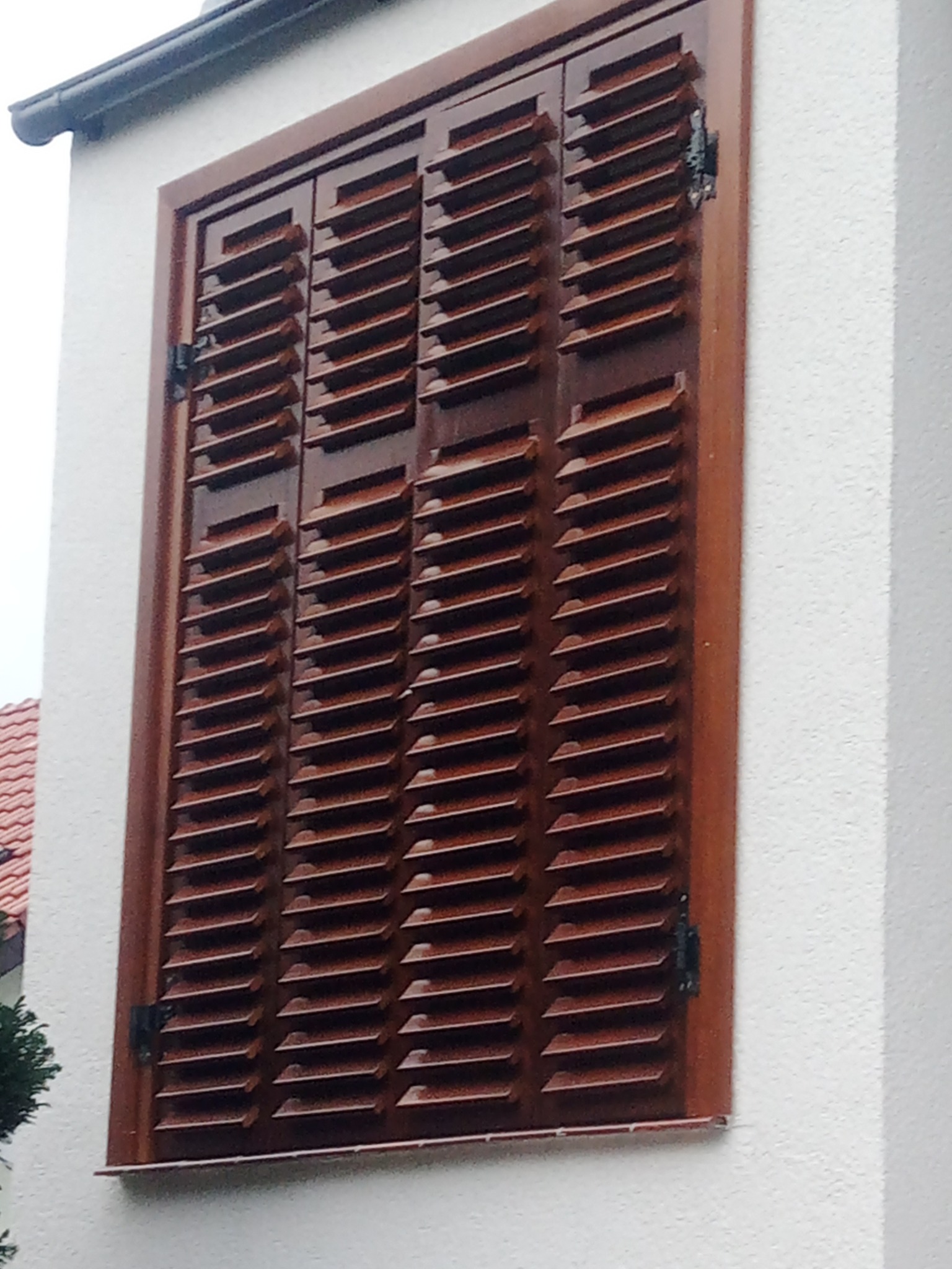 Zamknięte, brązowe, drewniane okiennice żaluzjowe zamontowane na białej elewacji budynku, widoczne czarne zawiasy.