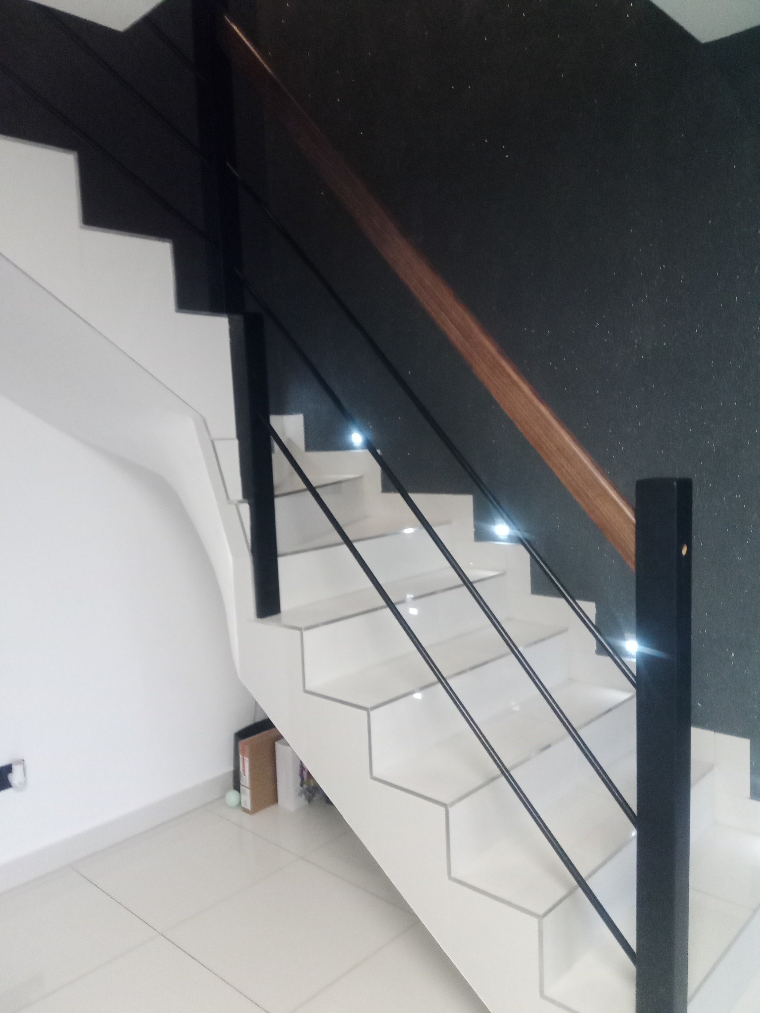 Nowoczesne schody drewniane z białymi stopniami o heksagonalnym kształcie, czarnymi balustradami i drewnianą poręczą, oświetlone punktowymi lampkami LED w stopniach, na tle ściany w kolorze...