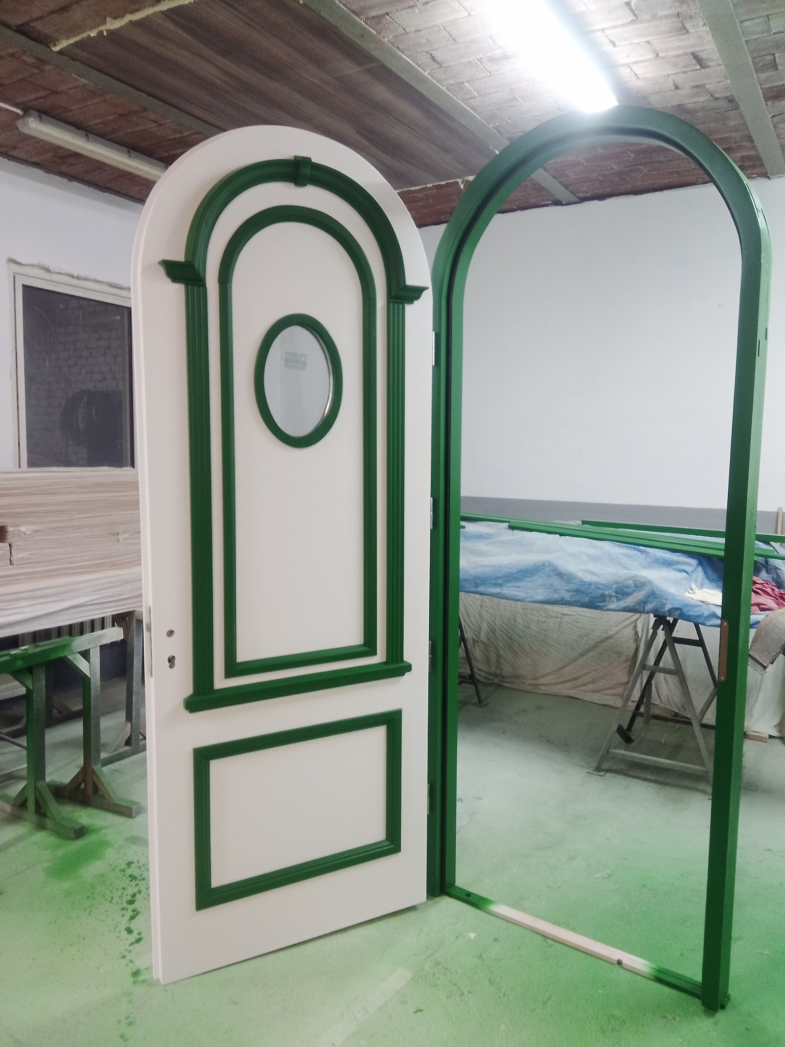 Białe drzwi wewnętrzne z zielonymi zdobieniami w stylu retro, otwarte i ustawione w ościeżnicy, widoczne ślady farby na podłodze, sugerujące etap wykończeniowy w warsztacie.