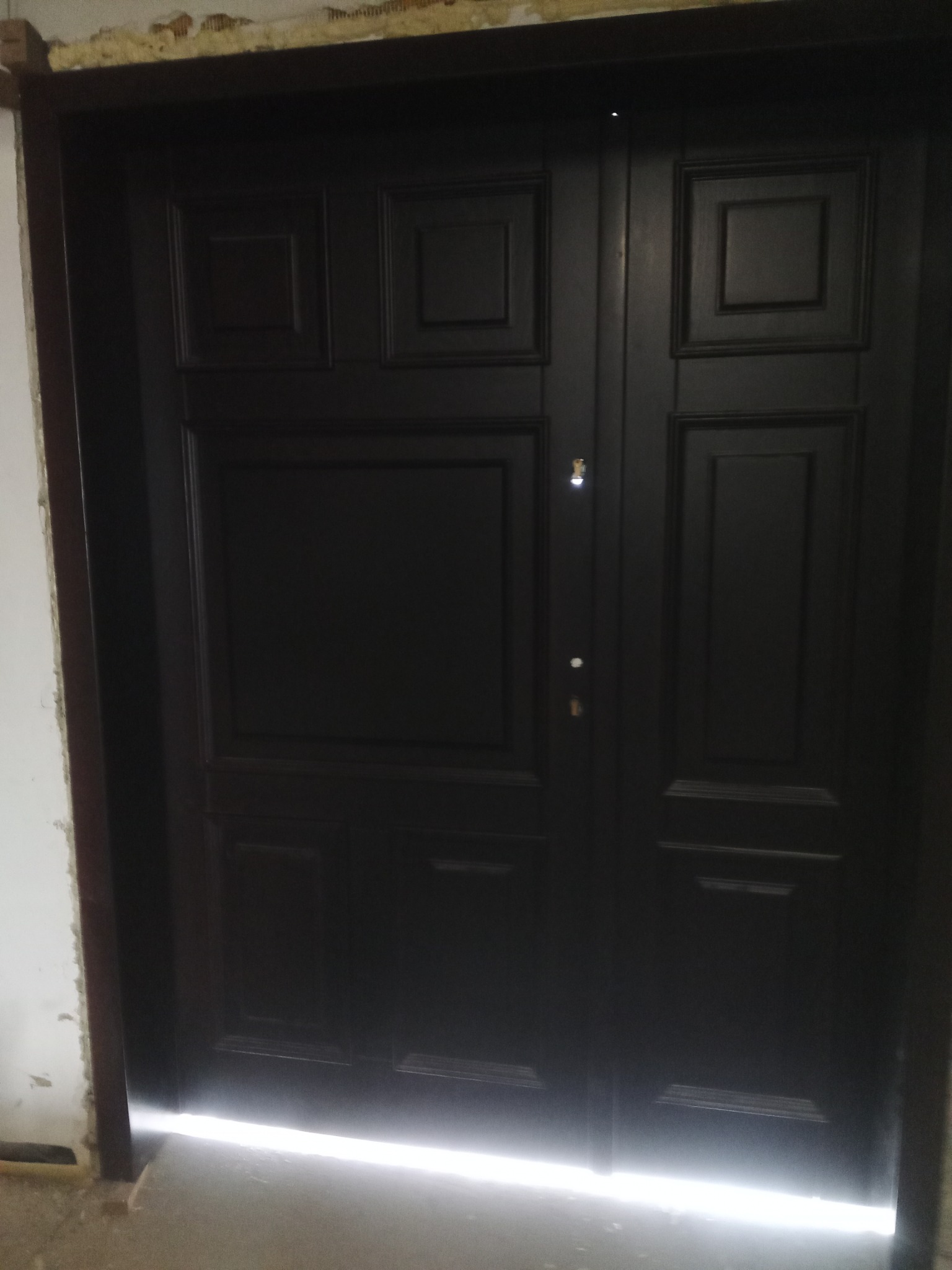 Ciemne, dwuskrzydłowe drzwi z panelami dekoracyjnymi, widoczne w trakcie montażu, ze szczeliną u dołu, przez którą wpada światło, ukazując surowy stan wykończenia podłogi i ścian.