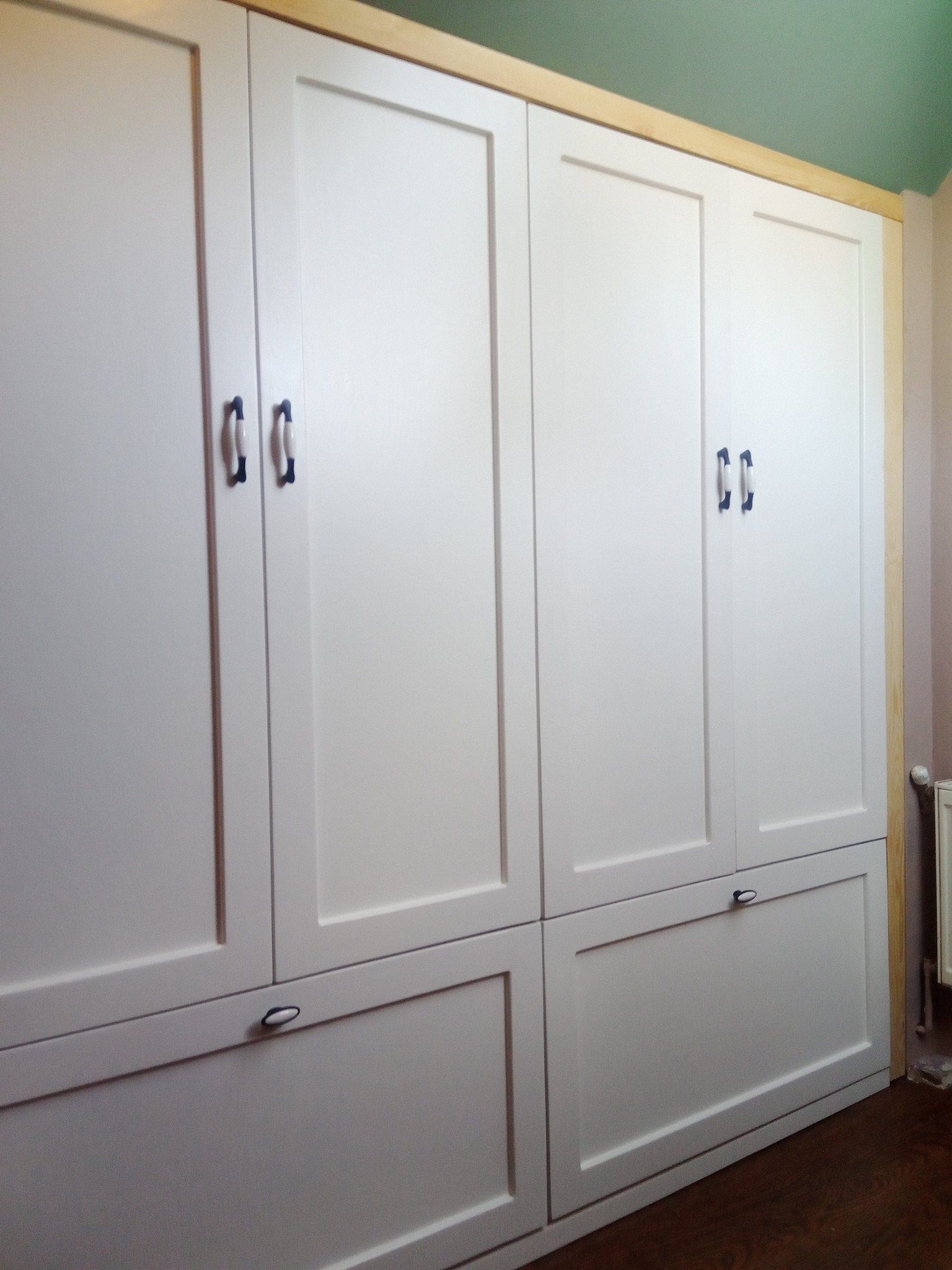 Biała, zabudowana szafa z drewnianymi frontami, ciemnymi uchwytami i widocznym fragmentem drewnianej ramy oraz podłogi.