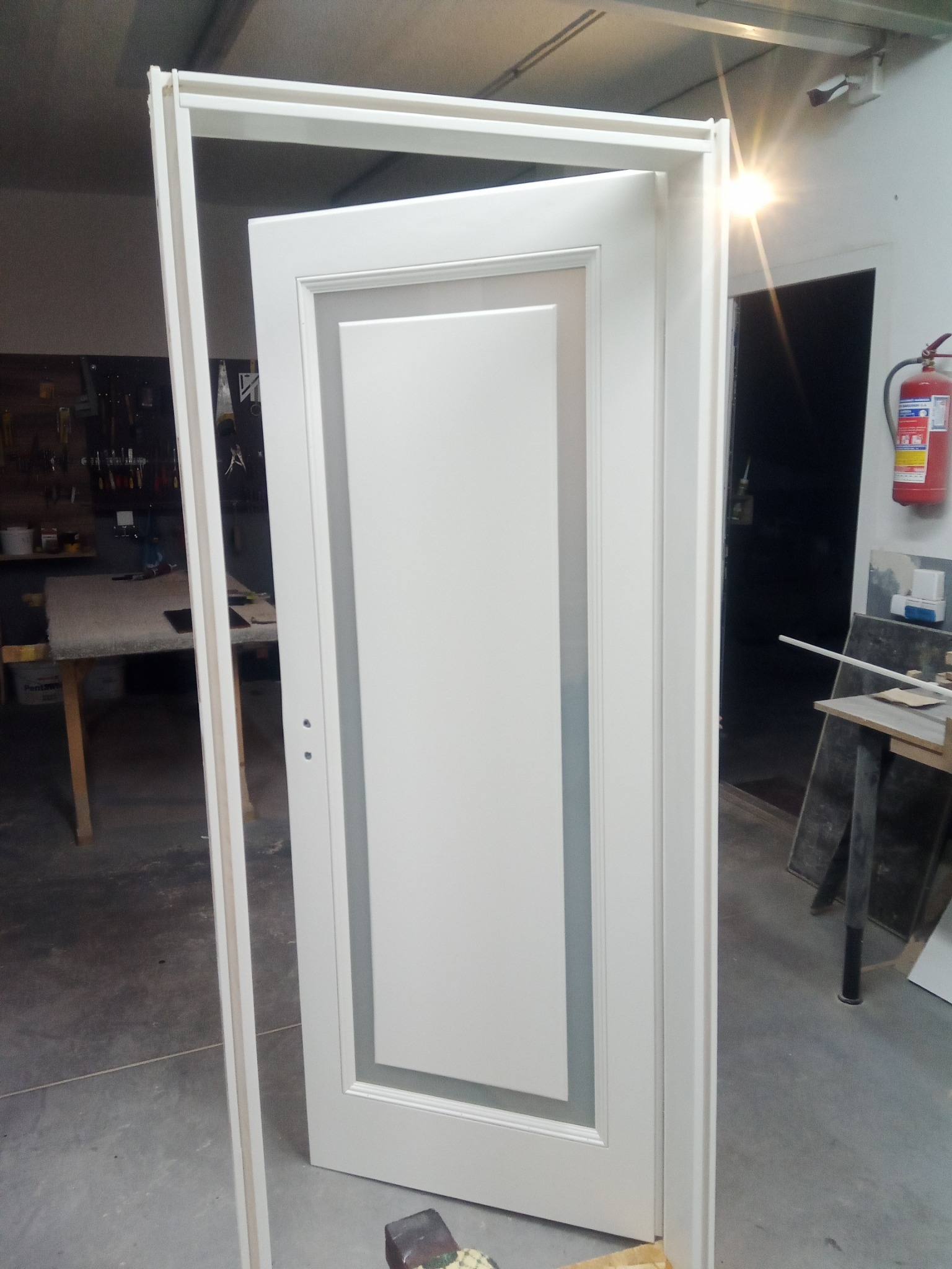 Białe, częściowo otwarte drzwi wewnętrzne z szarym obramowaniem w trakcie montażu ościeżnicy. Warsztatowy nieład w tle, widoczny sprzęt i narzędzia.