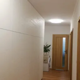 Długa, biała szafa na wymiar z drewnianym wzorem na frontach, zamontowana w korytarzu z drewnianą podłogą. W tle widoczna pralka i doniczka z rośliną.