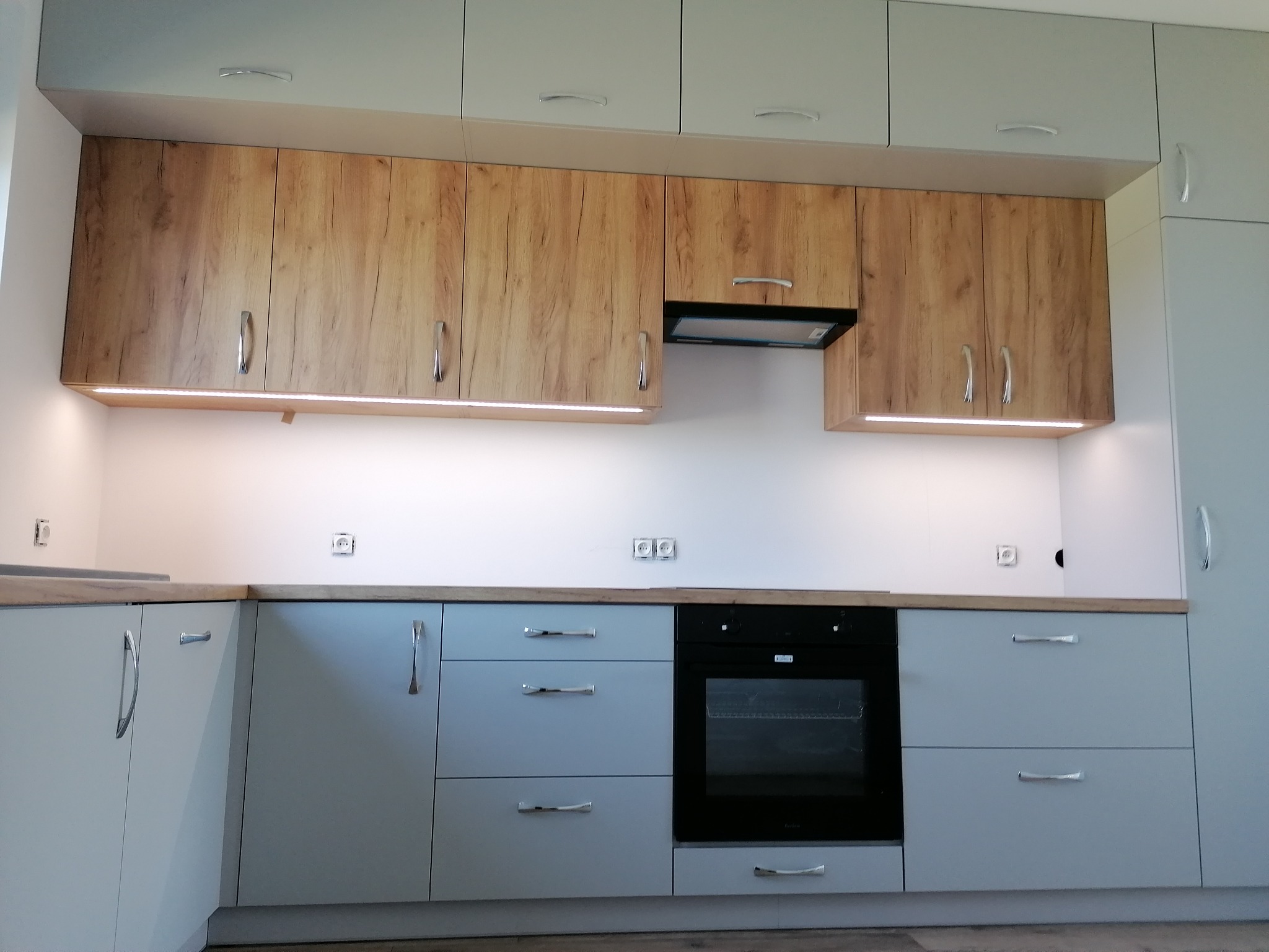 Jasnoszara zabudowa kuchenna z drewnopodobnymi górnymi szafkami i podświetleniem LED pod nimi, czarny piekarnik w zabudowie.
