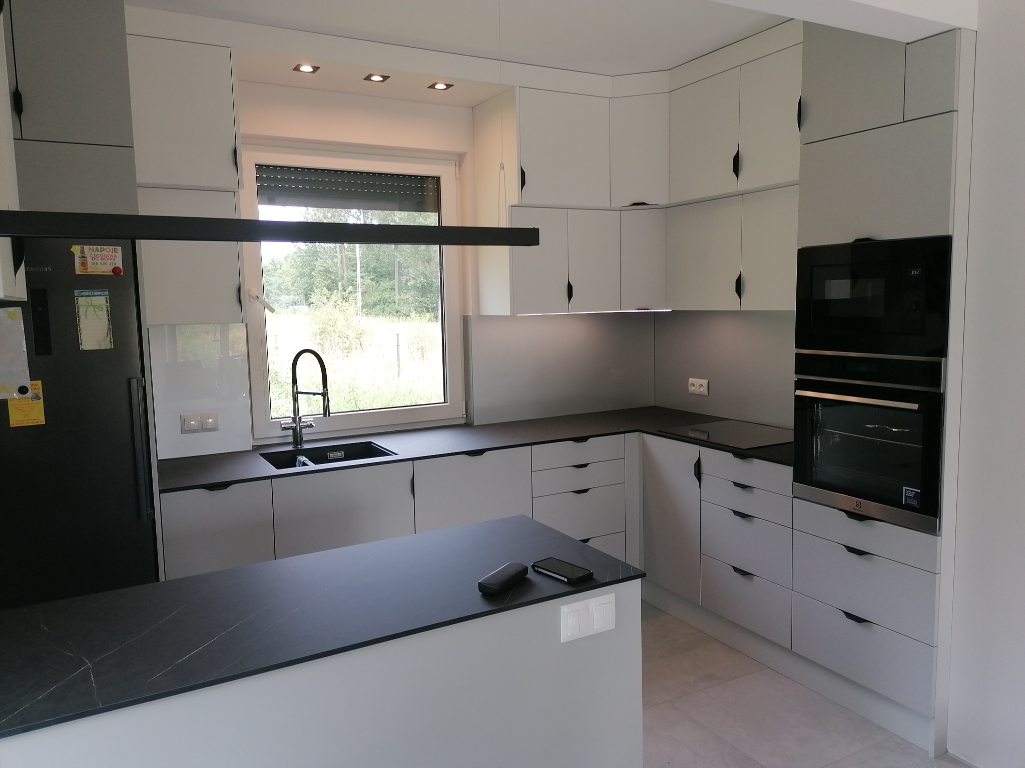 Nowoczesna kuchnia w odcieniach szarości z czarnym blatem i minimalistycznymi uchwytami szafek, wyspa kuchenna z czarnym blatem, widok na okno z roletami.