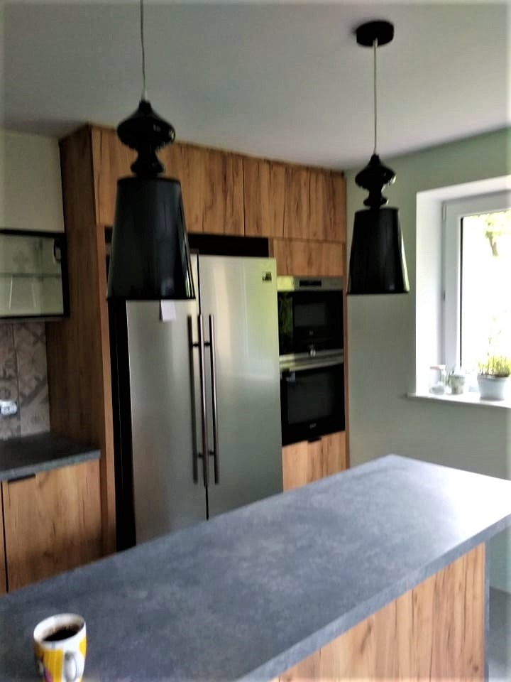 Nowoczesna kuchnia z drewnianymi frontami, wyspą z szarym blatem i czarnymi lampami wiszącymi; widoczny fragment okna z roślinami.