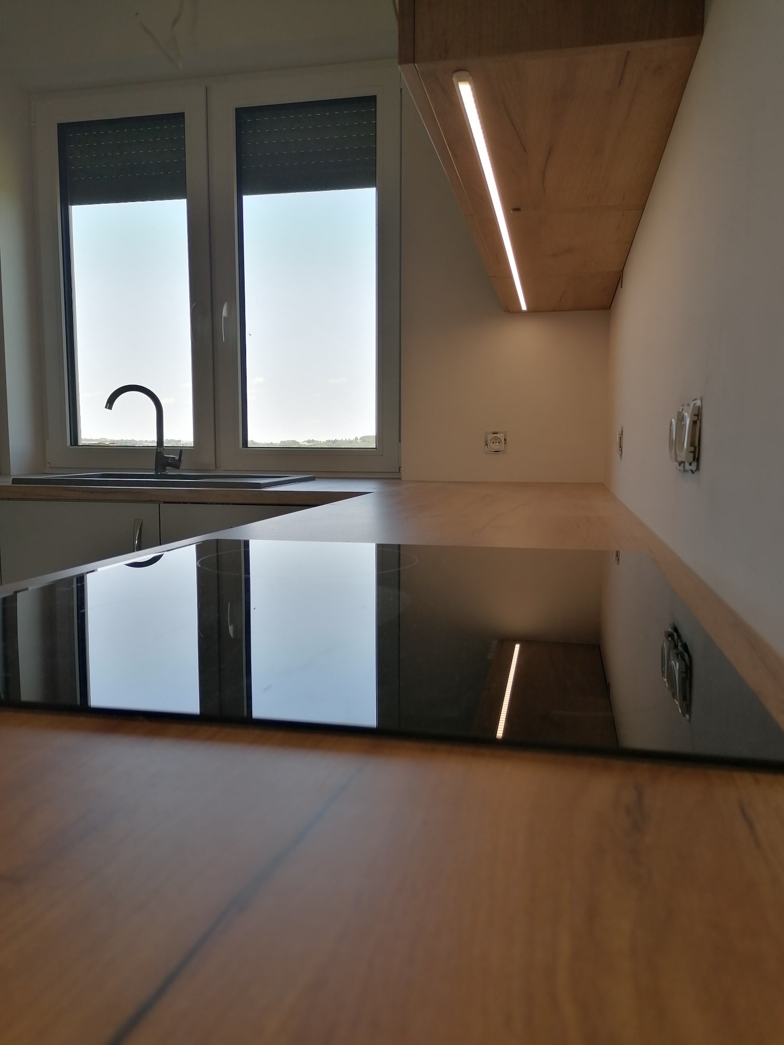 Nowoczesna kuchnia z blatem w kolorze drewna i czarną płytą indukcyjną, widok z perspektywy blatu, okno z roletami w tle, Sędziszów Małopolski.