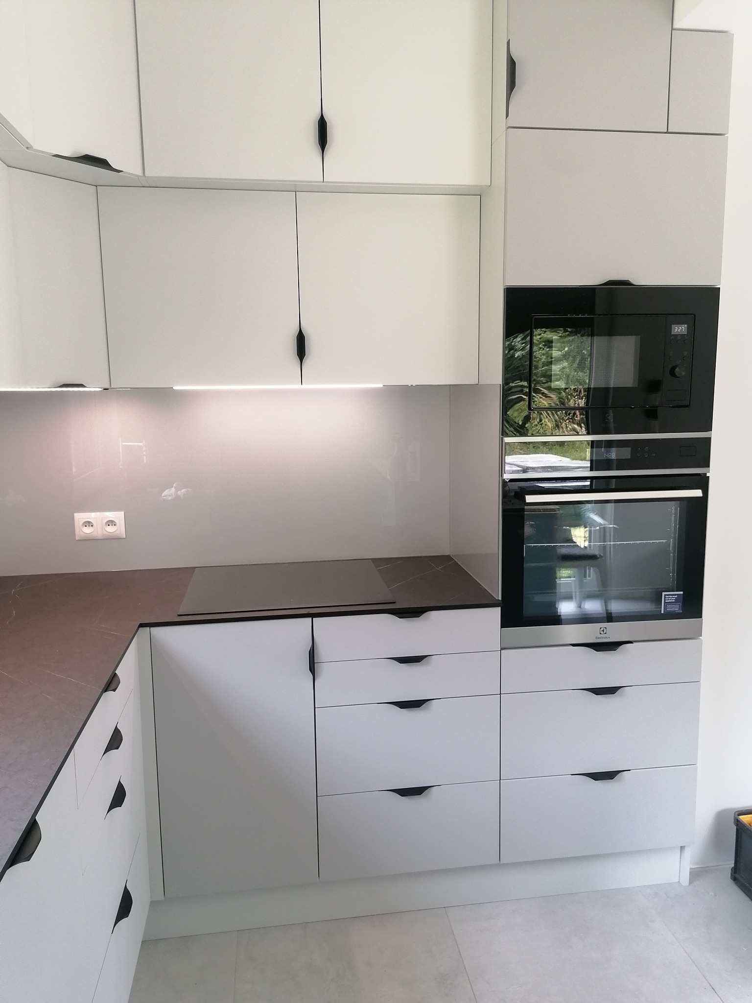 Nowoczesna, minimalistyczna kuchnia: białe szafki z czarnymi uchwytami, szary blat, płyta indukcyjna i zabudowane urządzenia AGD.