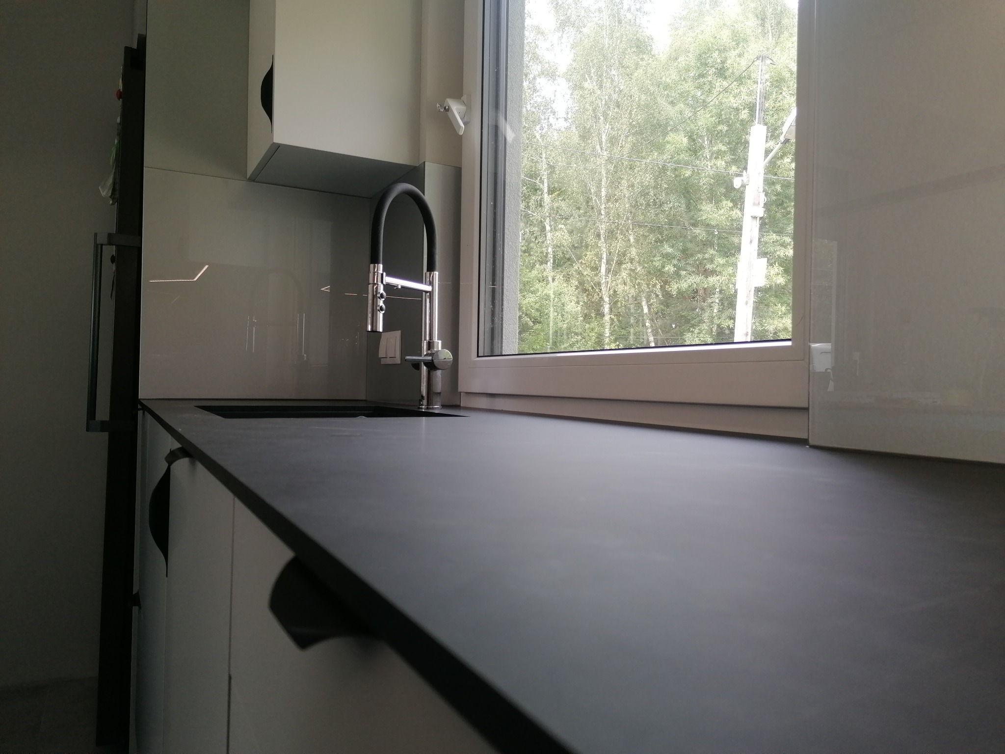 Nowoczesna kuchnia z czarnym blatem, chromowaną baterią i białymi szafkami, widok na zieleń za oknem.