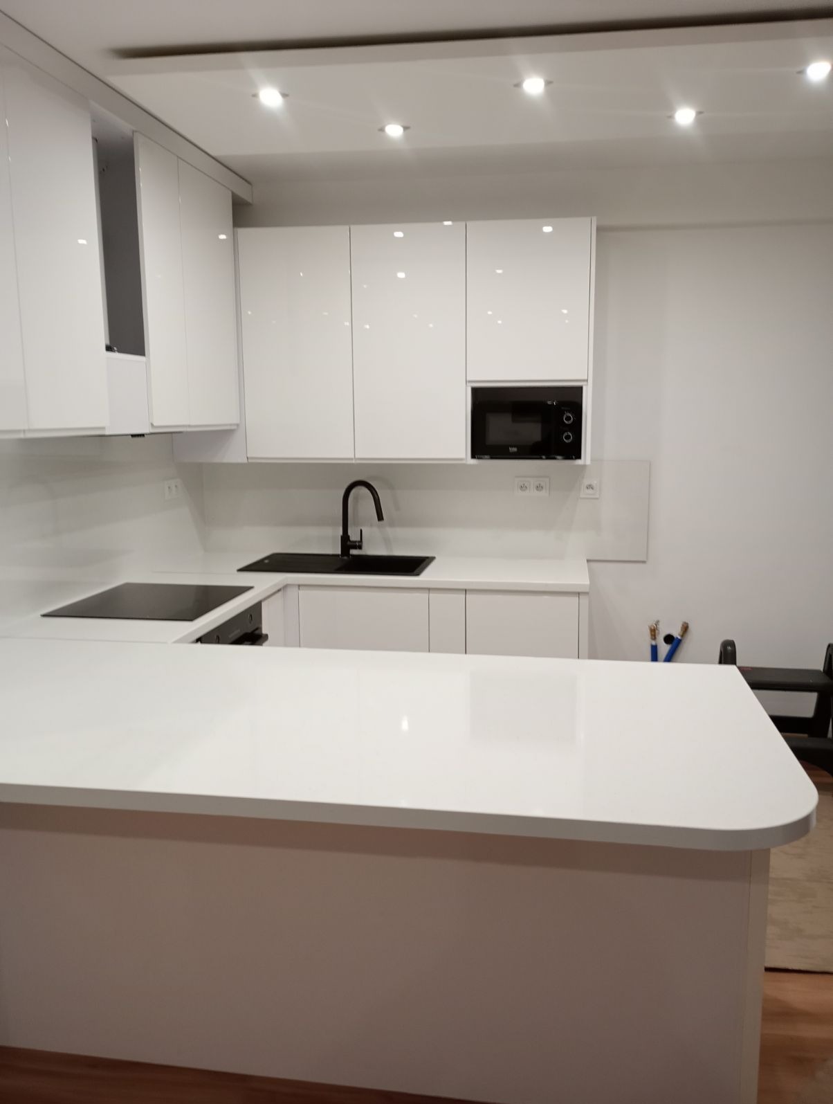 Nowoczesna, minimalistyczna kuchnia z białymi, lakierowanymi frontami, czarną armaturą i płytą indukcyjną, oświetleniem punktowym i blatem z jasnego konglomeratu.