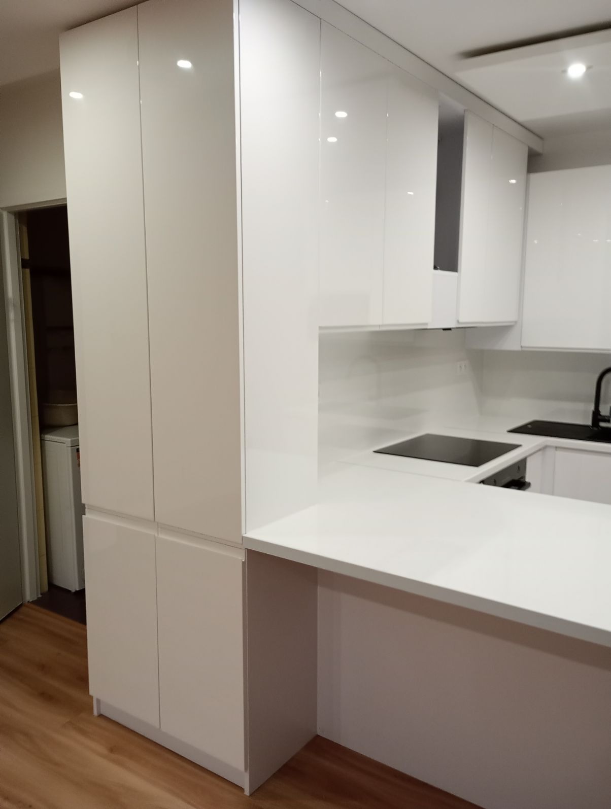 Biała, nowoczesna zabudowa kuchenna z wysokim połyskiem, obejmująca szafki stojące i wiszące, z blatem roboczym z konglomeratu i widoczną płytą indukcyjną.