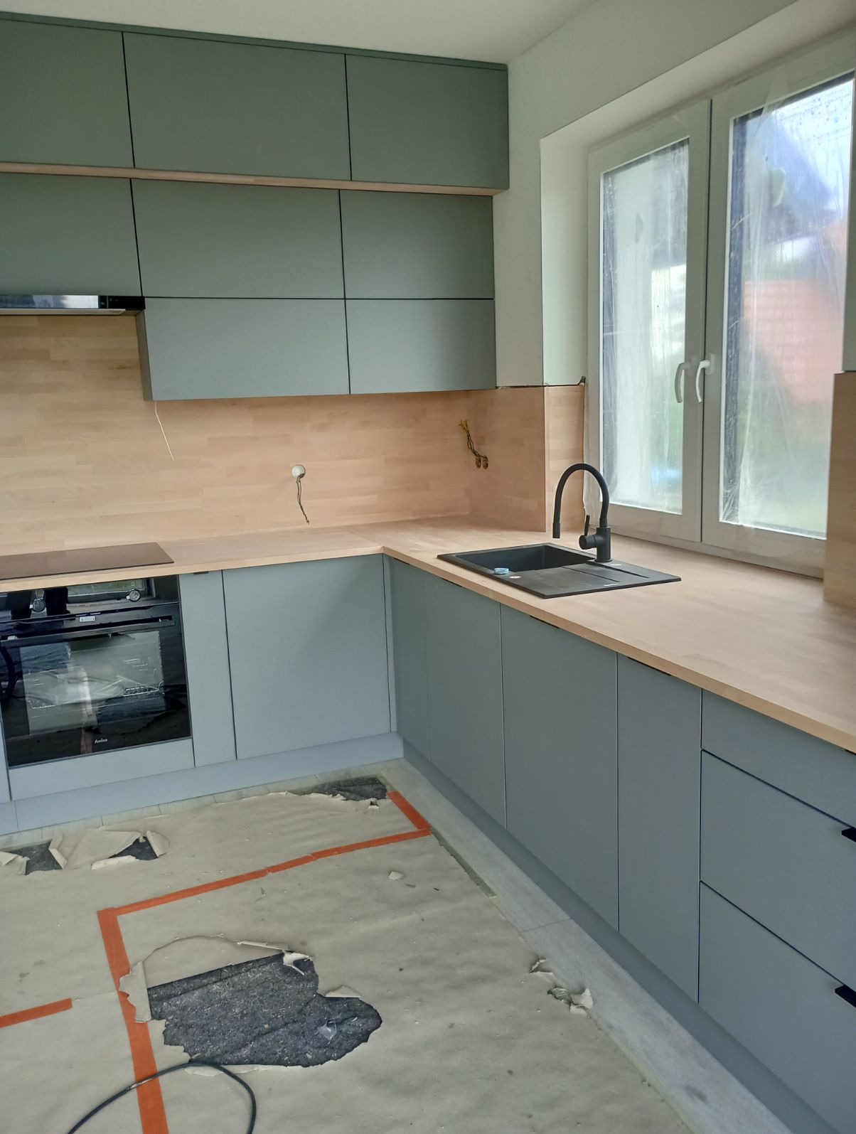 Szare szafki kuchenne z drewnianym blatem, czarnym zlewem i baterią, widok na okno, podłoga w trakcie remontu.