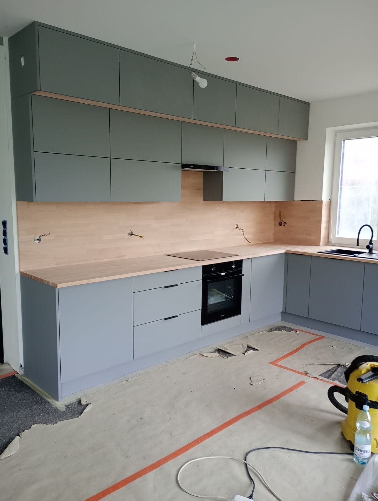 Montaż szarych szafek kuchennych z drewnianym blatem i panelem ściennym; widoczne przygotowania do podłączenia urządzeń i niedokończona podłoga zabezpieczona papierem malarskim.