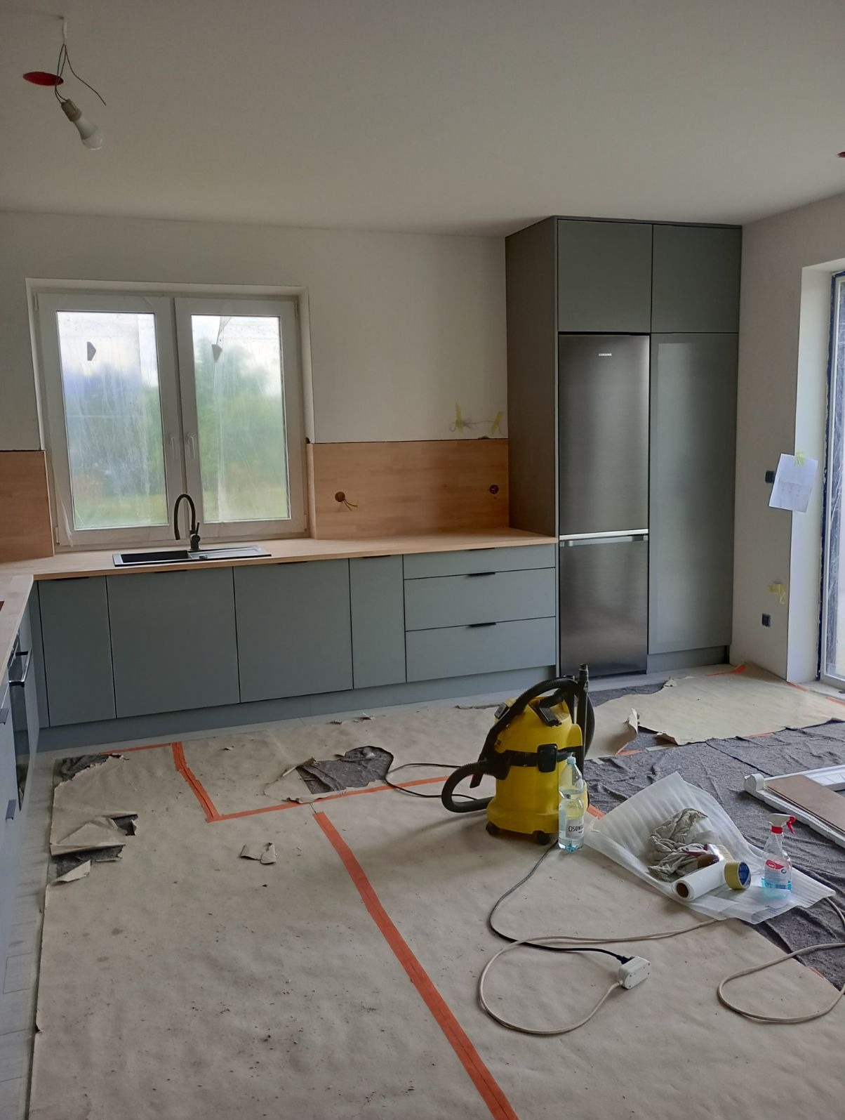 Montaż szarych mebli kuchennych z drewnianym blatem, widoczny fragment okna, lodówka i odkurzacz na podłodze zabezpieczonej folią malarską.