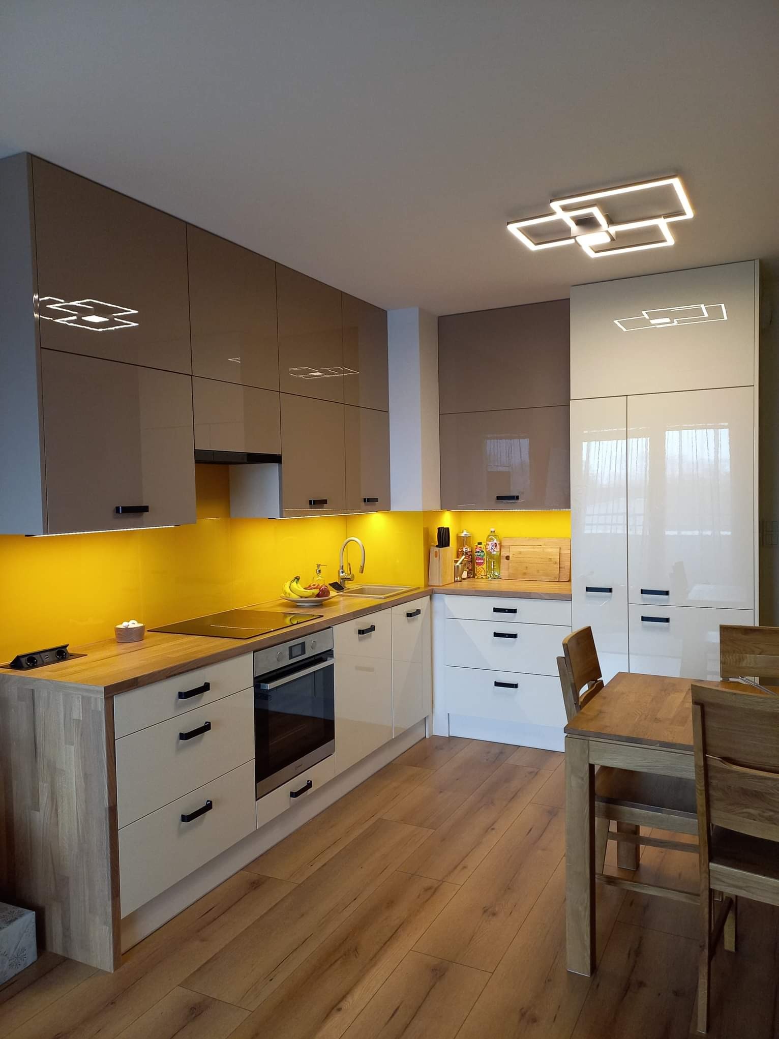 Nowoczesna kuchnia na wymiar z błyszczącymi frontami w odcieniach beżu i bieli, podświetlona żółtym szkłem lacobel, drewniany stół z krzesłami w tle.
