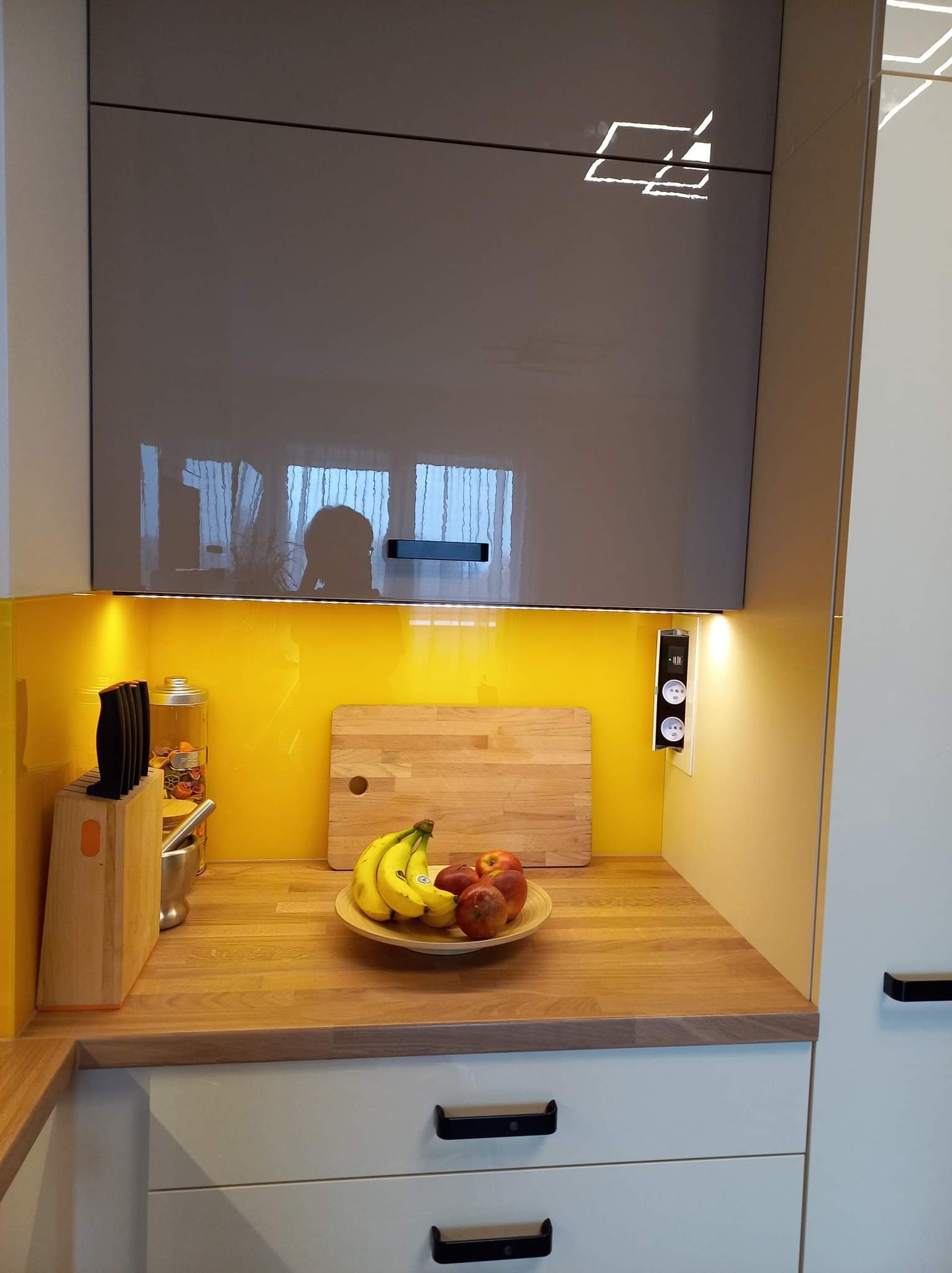 Fragment nowoczesnej kuchni z żółtym panelem, drewnianym blatem, na którym leżą owoce w misce, widoczne gniazdka elektryczne i oświetlenie LED pod szafkami.
