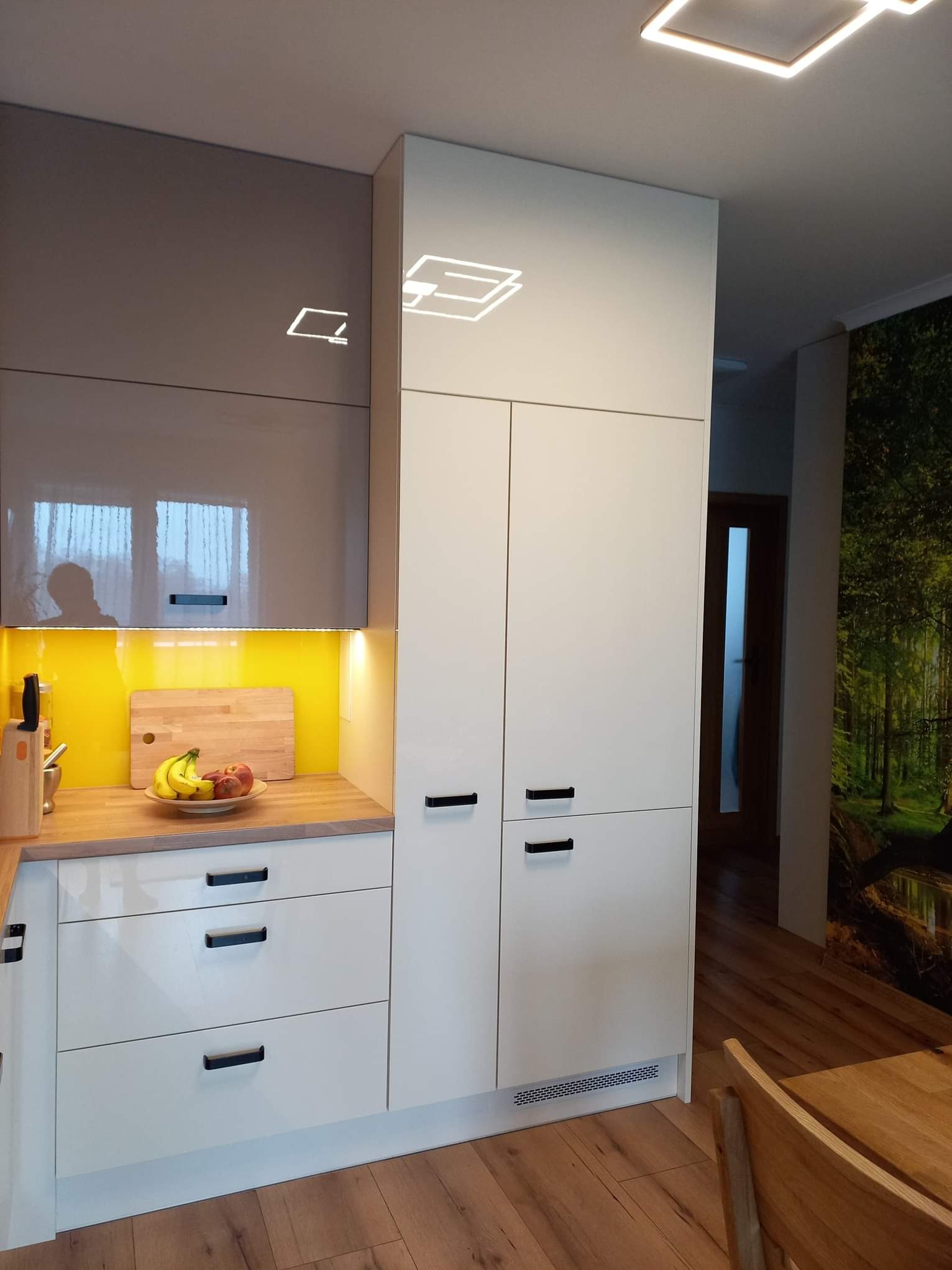 Nowoczesna zabudowa kuchenna z białymi frontami, drewnianym blatem i żółtym panelem ściennym, oświetlenie LED pod szafkami, na blacie owoce w misce i deska do krojenia, w tle fototapeta z motywem...