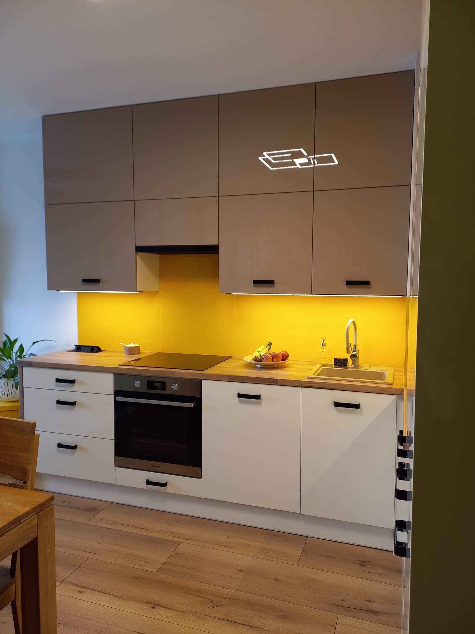 Nowoczesna kuchnia z beżowymi szafkami górnymi i białymi dolnymi, podświetlona żółtym światłem, z drewnianym blatem i podłogą, widoczne owoce i roślina doniczkowa.