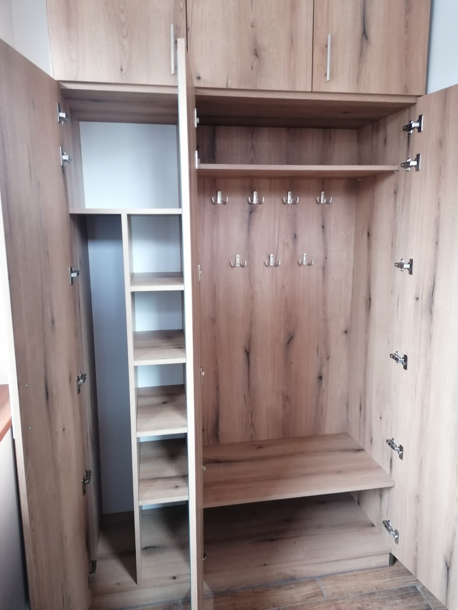 Otwarta szafa wnękowa z jasnego drewna z półkami i wieszakami, widoczne zawiasy i podział na strefy.