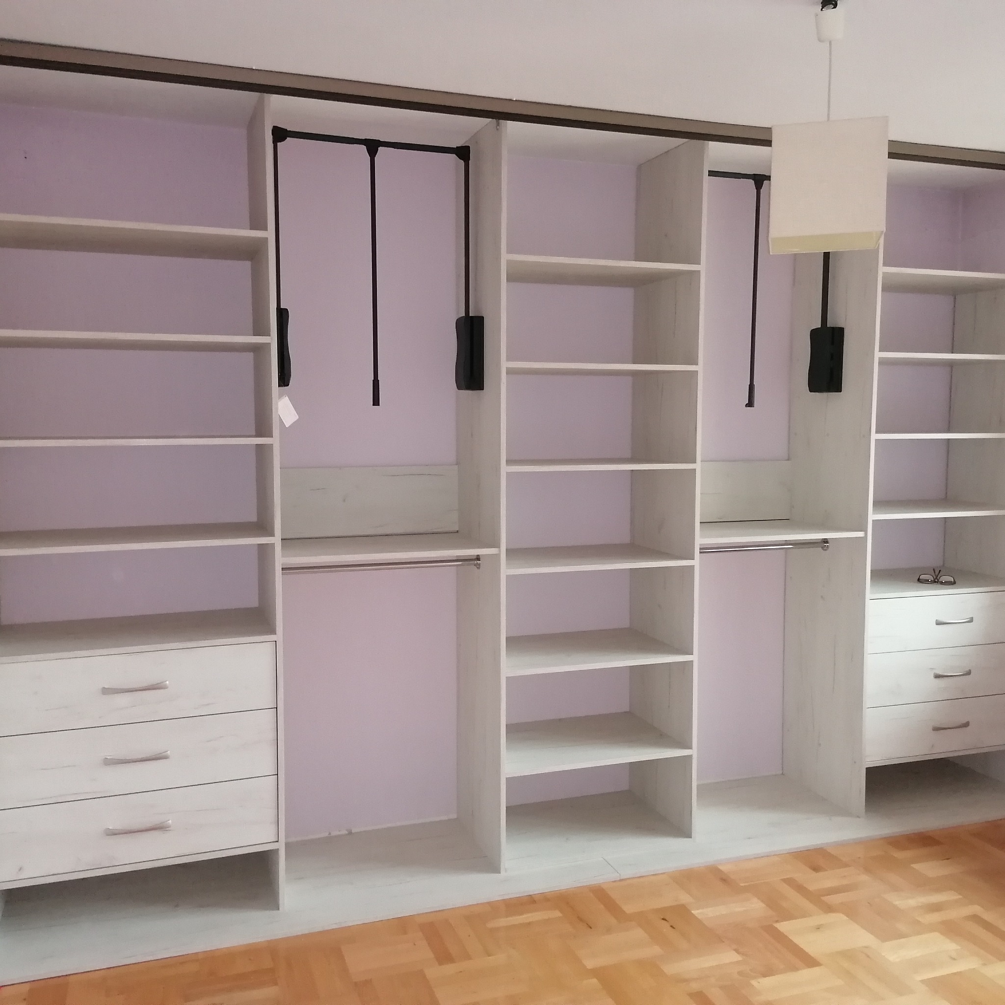 Jasna, otwarta garderoba z szufladami i półkami, wyposażona w drążki na ubrania, z drewnianą podłogą w tle.