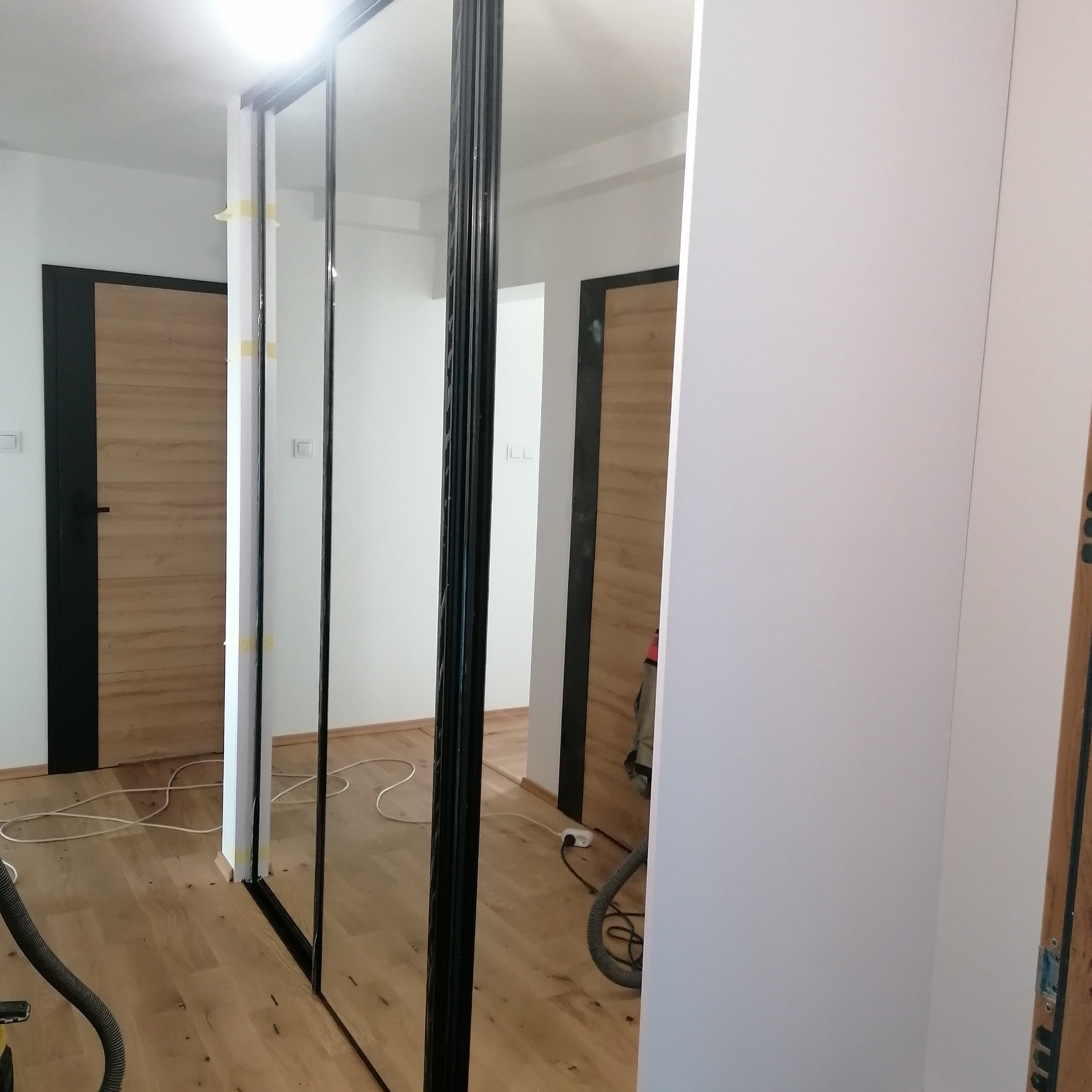 Montaż szafy wnękowej z lustrzanymi drzwiami przesuwnymi, widoczne taśmy malarskie zabezpieczające oraz przewody i odkurzacz na podłodze.