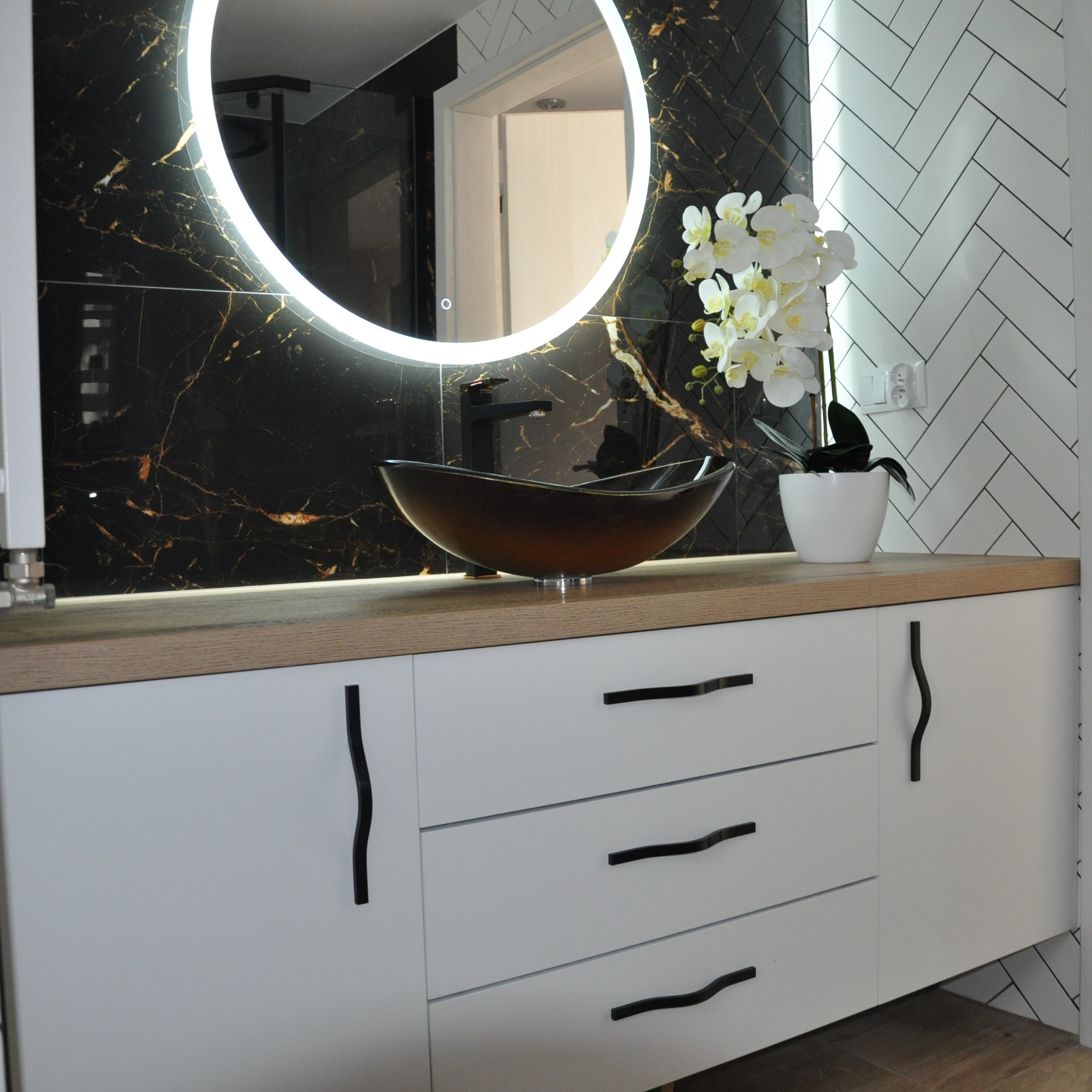 Nowoczesna łazienka z czarnym marmurem na ścianie, okrągłym lustrem z podświetleniem LED, brązową umywalką na blacie z drewnopodobnym dekorem i białymi szafkami z oryginalnymi, falistymi uchwytami.
