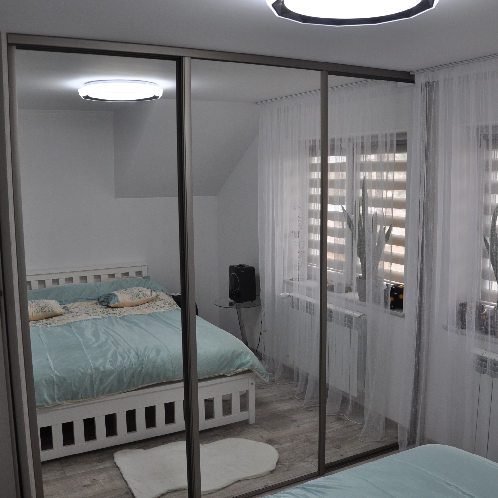 Szafa wnękowa z lustrzanymi przesuwnymi drzwiami, odbijająca fragment sypialni z białym łóżkiem i zasłonami w oknie.