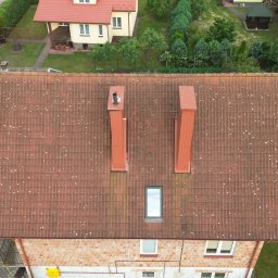 TWINSBRO SPÓŁKA Z OGRANICZONĄ ODPOWIEDZIALNOŚCIĄ - Widok z lotu ptaka na dach pokryty czerwoną dachówką z dwoma kominami i oknem dachowym, widoczne białe plamy mchu i porostów, w tle bujna zieleń drzew i trawników.