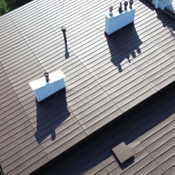 TWINSBRO SPÓŁKA Z OGRANICZONĄ ODPOWIEDZIALNOŚCIĄ - Brązowy dach z blachy na domu, widok z góry, z białymi kominami i elementami wykończeniowymi. Cień drzew na dachu. Regularny wzór blachy.