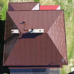 Widok z góry na brązowy dach domu z dwoma kominami, otoczony zielenią. Fragment dachu sąsiedniego budynku w tle. Dach po renowacji.