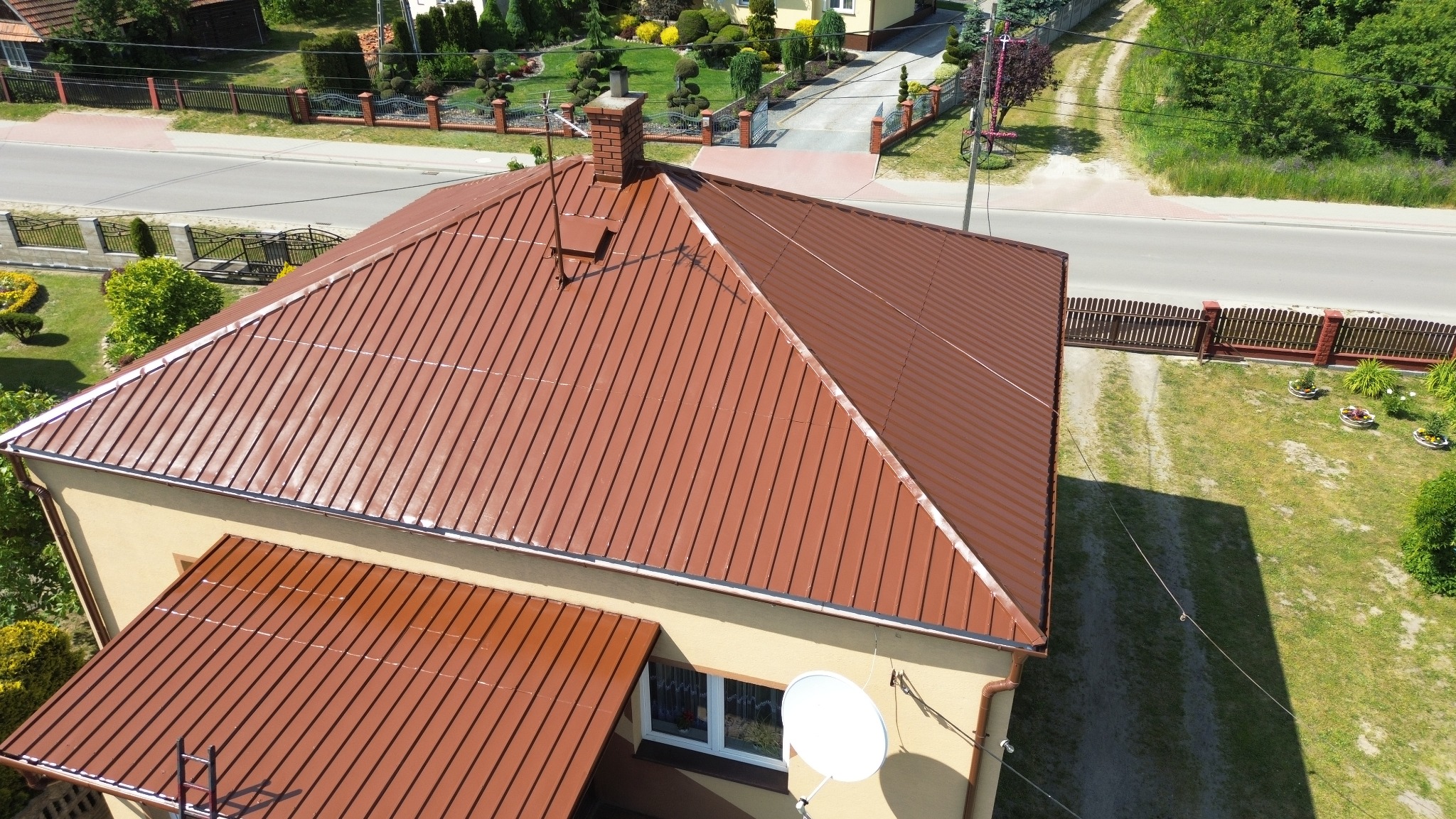 Dom z nowym, brązowym dachem z blachy trapezowej, widok z góry, z anteną satelitarną na elewacji i starannie utrzymanym ogrodem w tle.