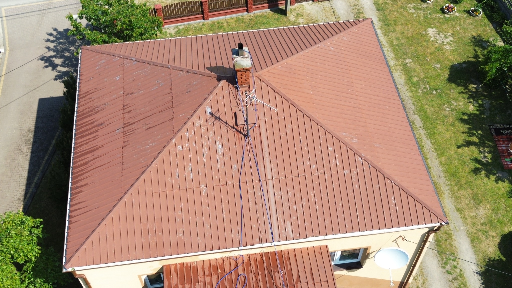 Widok z góry na brązowy dach z blachy trapezowej z ceglanym kominem i anteną satelitarną, częściowo porośnięty mchem, z widocznymi śladami zabrudzeń i zacieków.