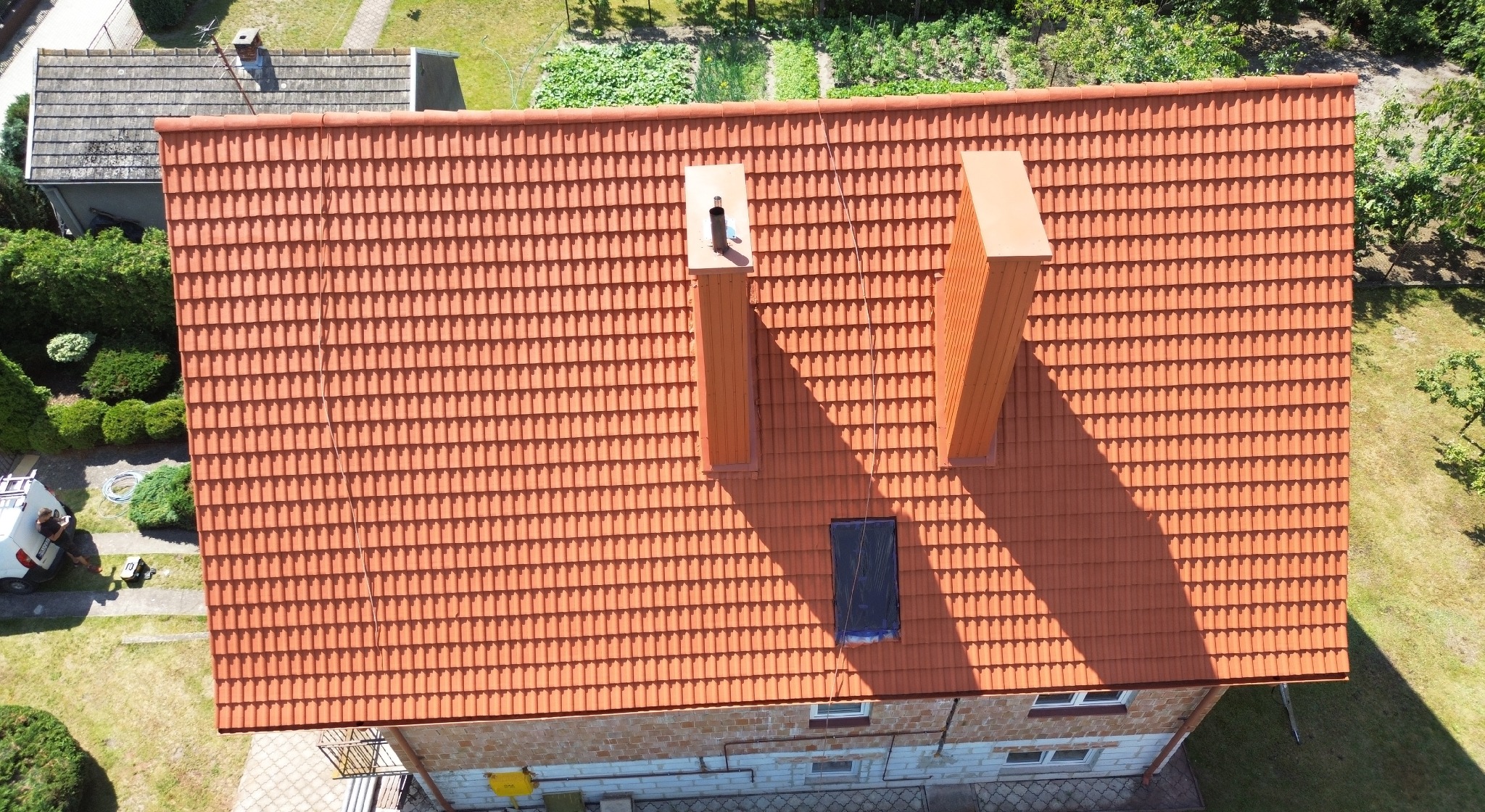 Widok z góry na dach pokryty czerwoną dachówką ceramiczną, z dwoma kominami i zabezpieczonym oknem dachowym, otoczony zielenią i częściowo widocznym samochodem dostawczym.