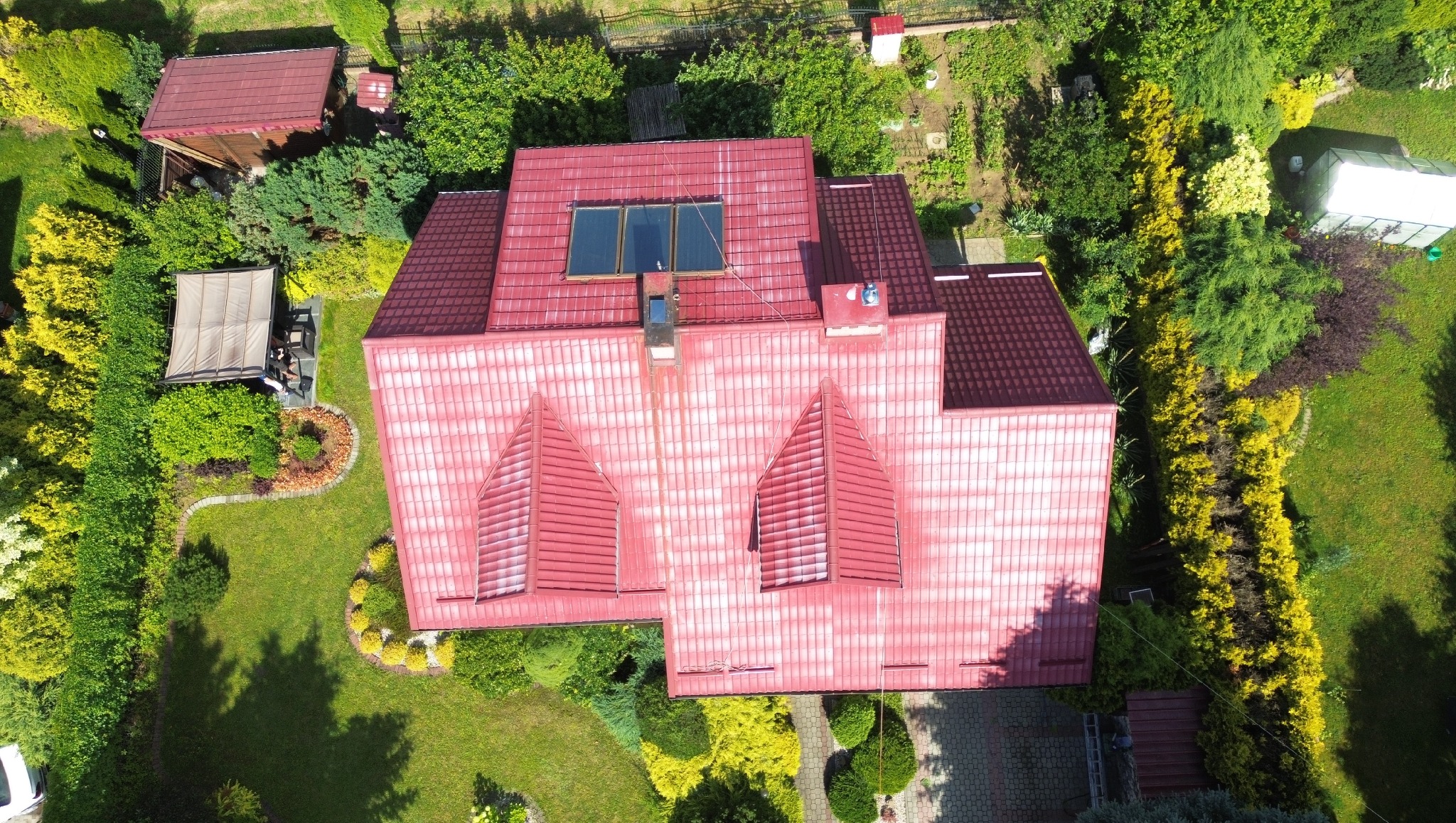 Widok z lotu ptaka na dom z czerwoną dachówką, na której widać ślady czyszczenia, z oknami dachowymi i kominem, otoczony bujnym ogrodem z altaną i drzewami.