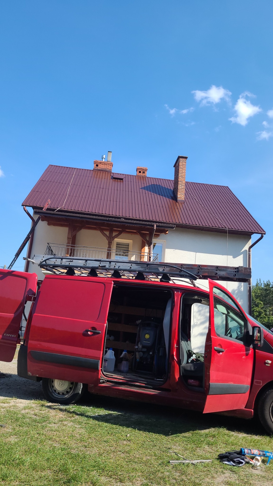 Czerwony bus z otwartymi drzwiami na tle domu z nowym, brązowym dachem i niebieskiego nieba. Na dachu widoczne kominy. Na dachu oparta drabina.