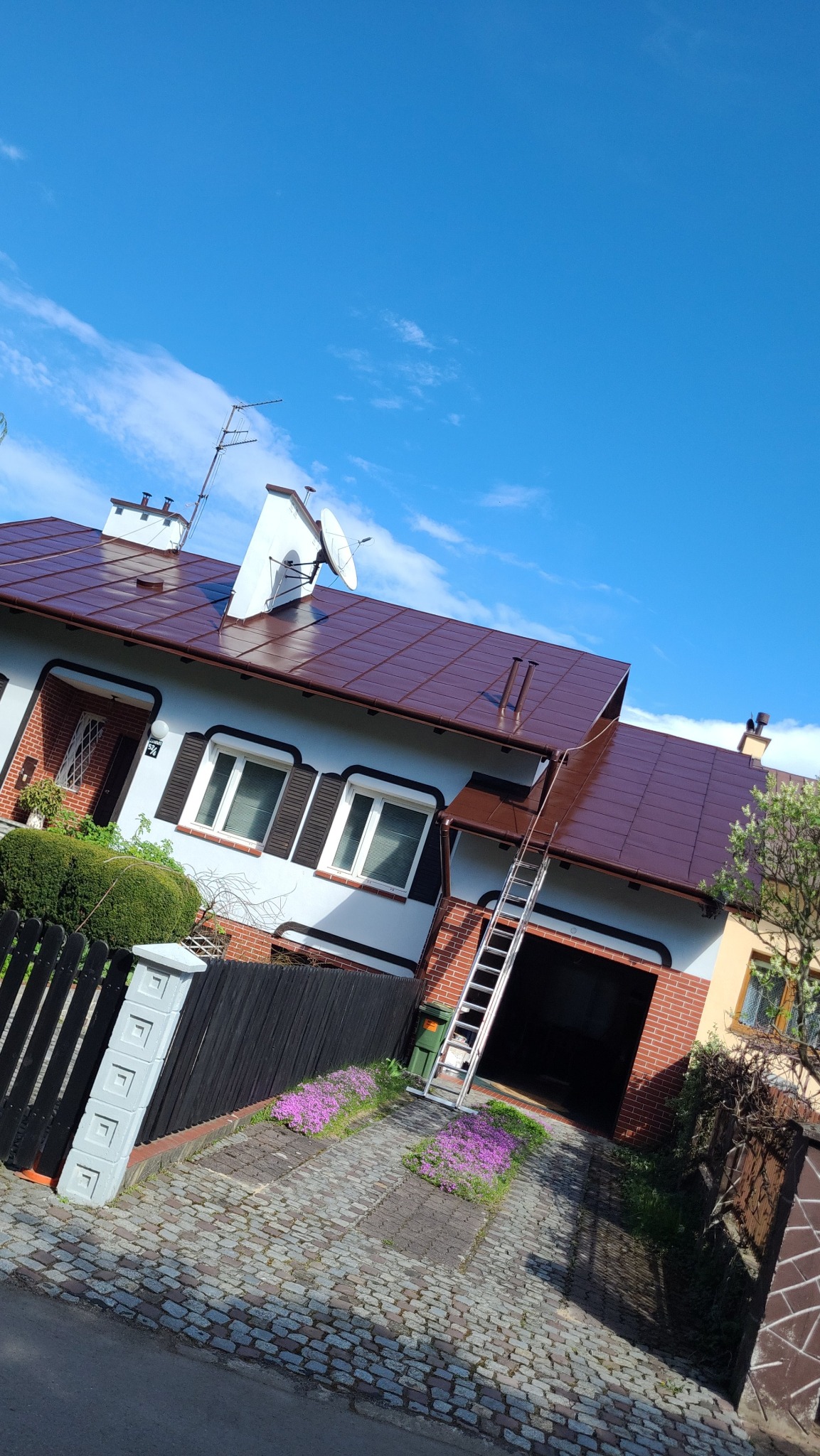 Dom jednorodzinny z nowym, brązowym dachem, częściowo ocieploną elewacją i przystawioną drabiną. Widok z dołu, błękitne niebo w tle.