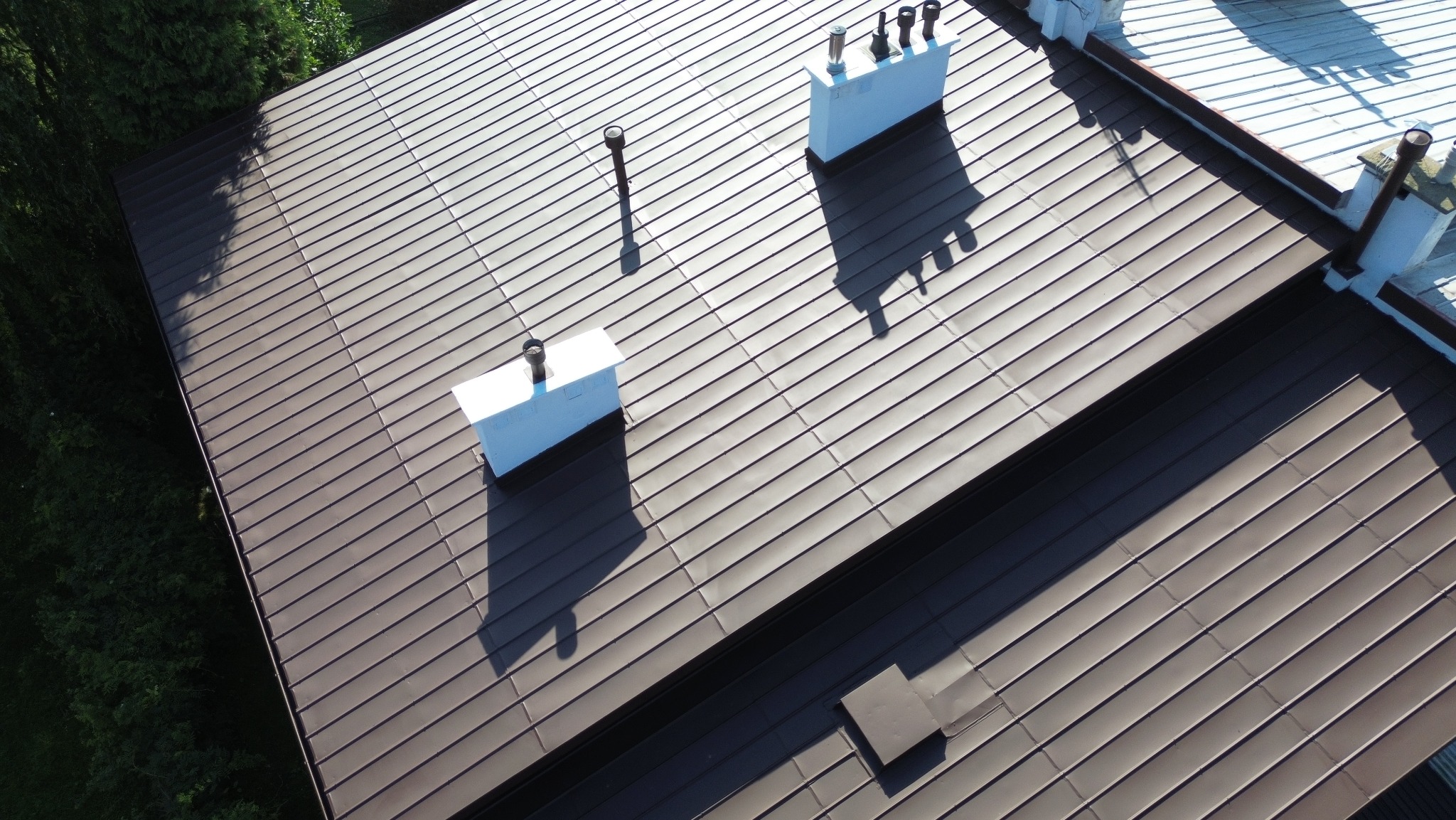 Brązowy dach z blachy na domu, widok z góry, z białymi kominami i elementami wykończeniowymi. Cień drzew na dachu. Regularny wzór blachy.