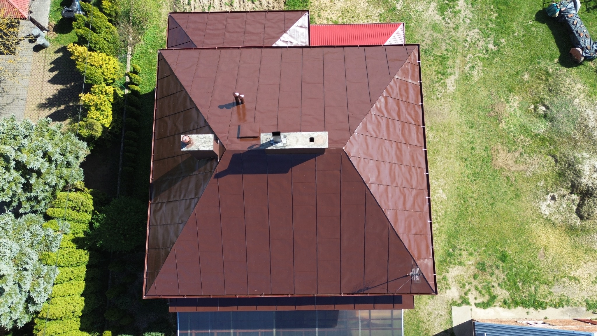 Widok z góry na brązowy dach domu z dwoma kominami, otoczony zielenią. Fragment dachu sąsiedniego budynku w tle. Dach po renowacji.