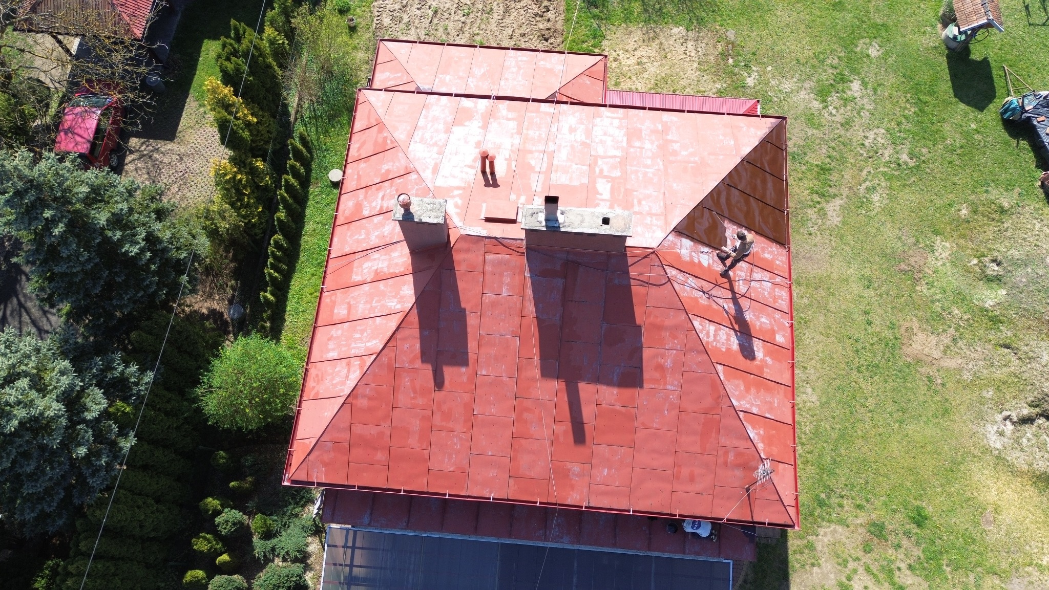Czerwony dach z blachy widziany z góry, z kominami i osobą pracującą na jego powierzchni, otoczony zielenią. Widoczne ślady po czyszczeniu dachu.