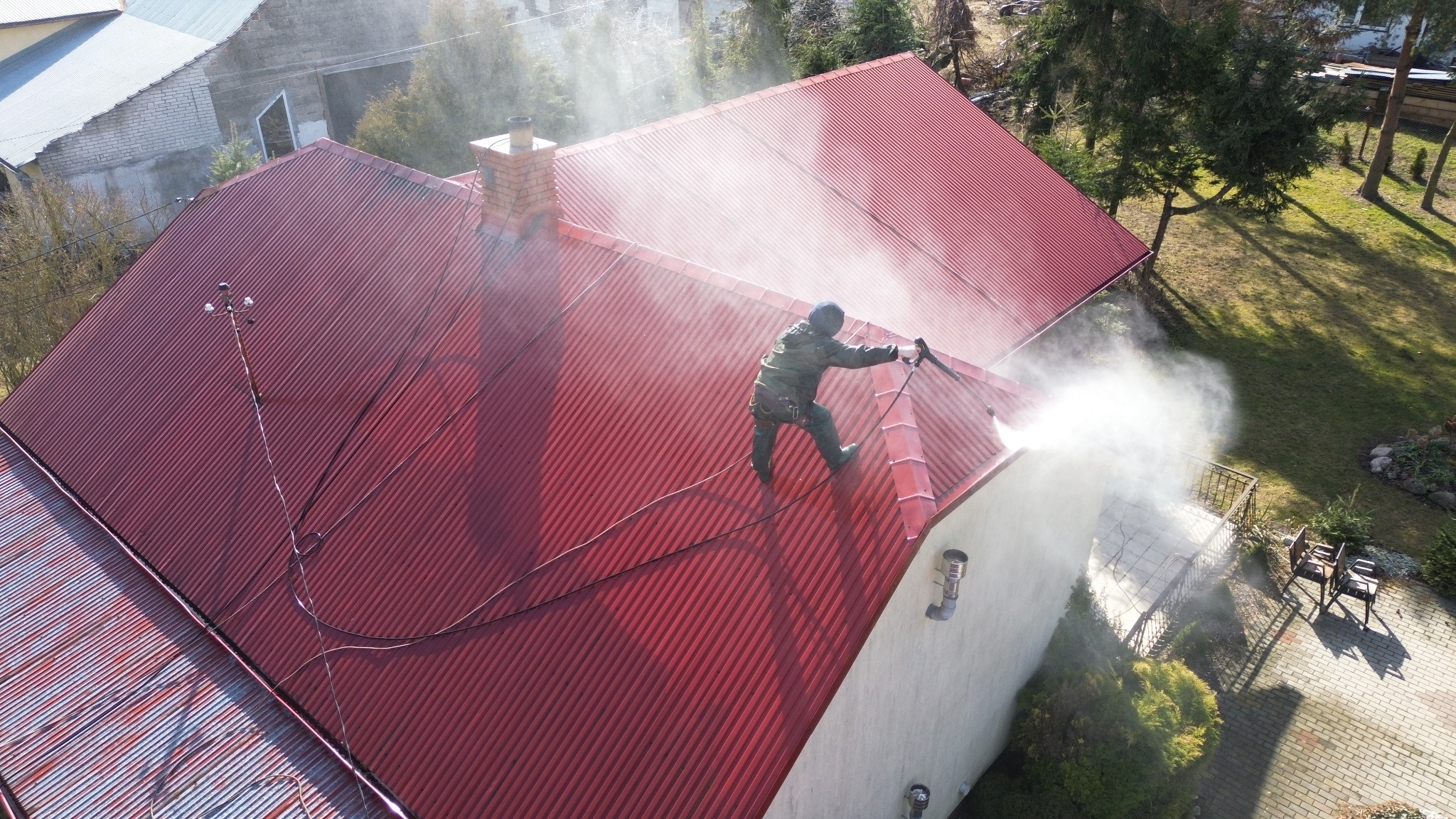 Czerwony dach z blachy trapezowej czyszczony myjką ciśnieniową przez pracownika w kombinezonie, widok z góry na dom z ceglanym kominem i zielonym ogrodem.