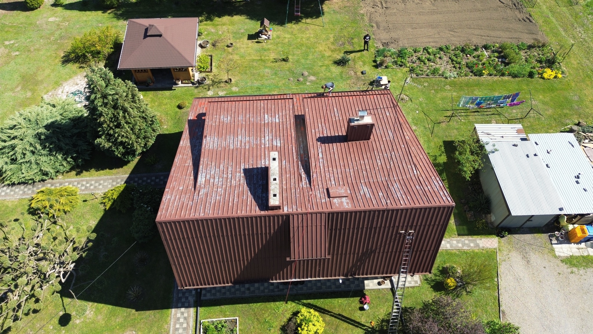 Widok z góry na duży, brązowy dach z blachy falistej z dwoma kominami. Na dachu pracują osoby. Wokół zielony trawnik i zabudowania gospodarcze. Widoczna drabina oparta o ścianę budynku.