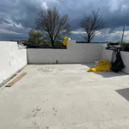 Wnętrze fundamentów domu murowanego z bloczków betonowych, zabezpieczone żółtą folią, widok na pochmurne niebo i drzewa w tle.