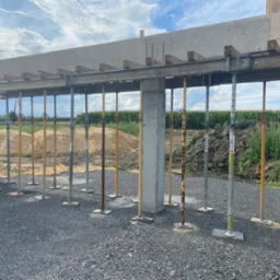 Wznoszenie betonowego nadproża z użyciem metalowych podpór teleskopowych na placu budowy domu murowanego, widoczne pustaki i krajobraz polny w tle.