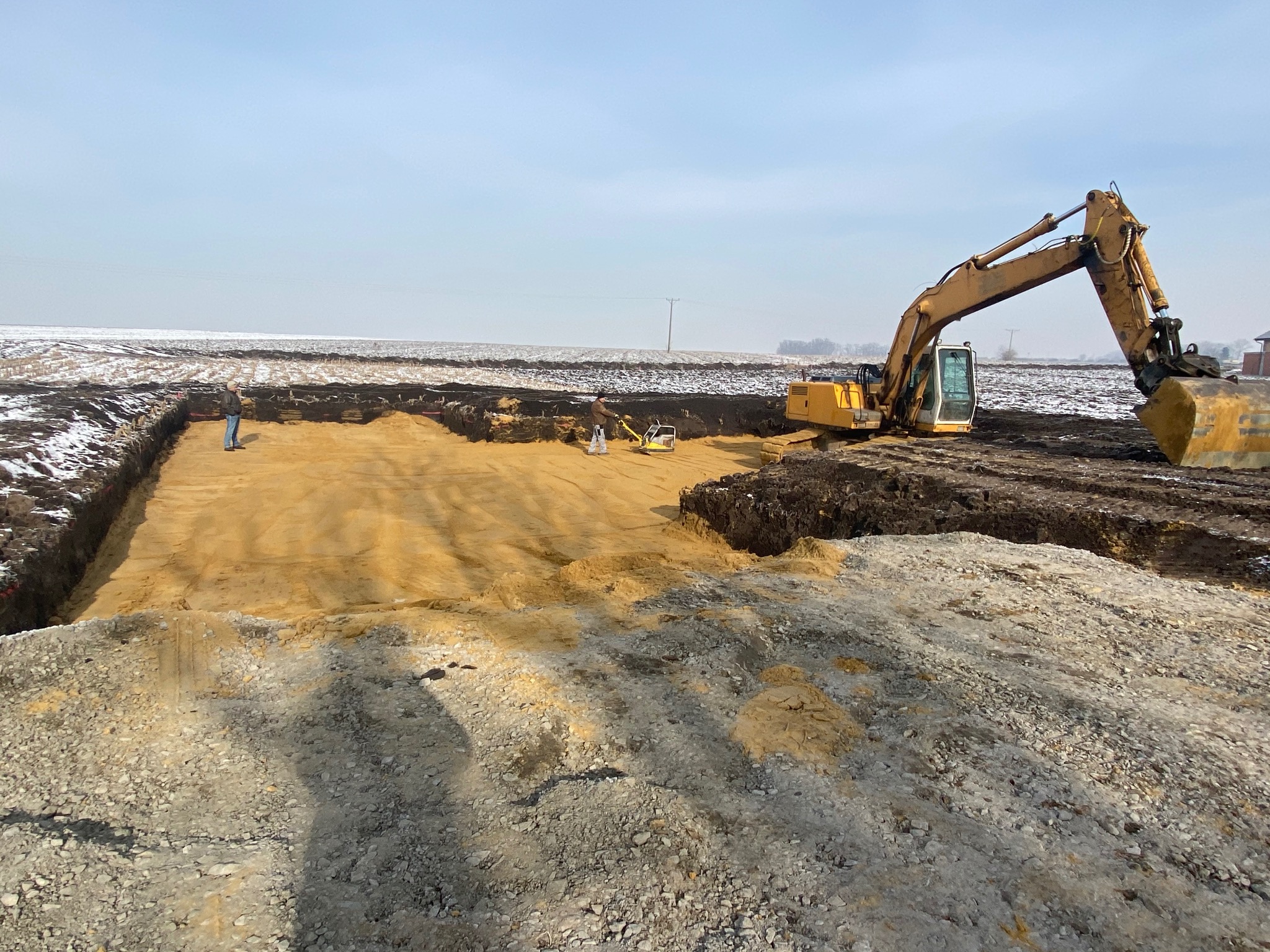 Żółta koparka na placu budowy, w tle pracownik i ubijarka gruntu, ziemia częściowo pokryta śniegiem.