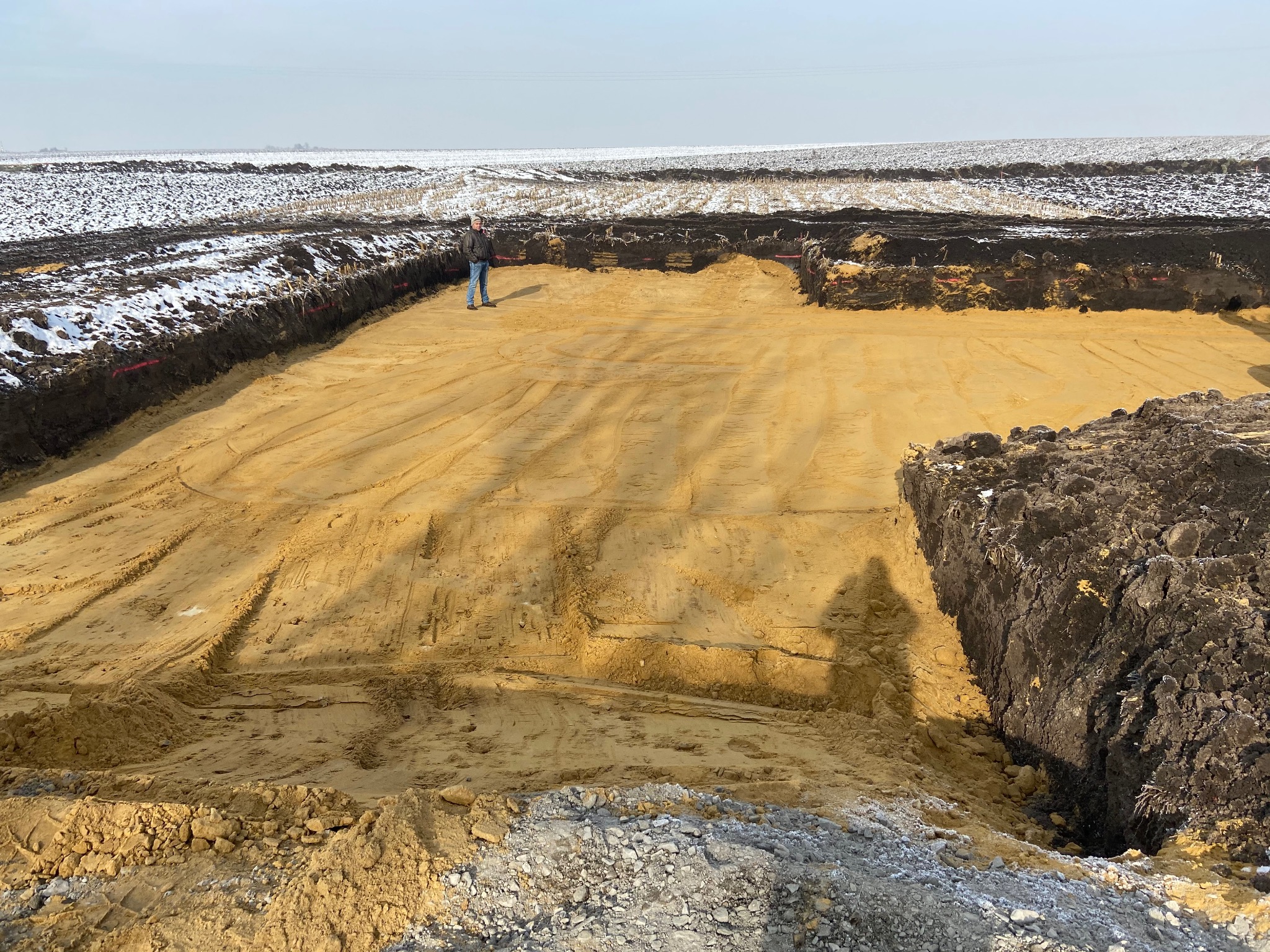 Szeroki wykop pod budynek na piaszczystym terenie, z widoczną osobą w oddali dla skali, otoczony ziemią i częściowo pokryty śniegiem, wykonany w pochmurny dzień.