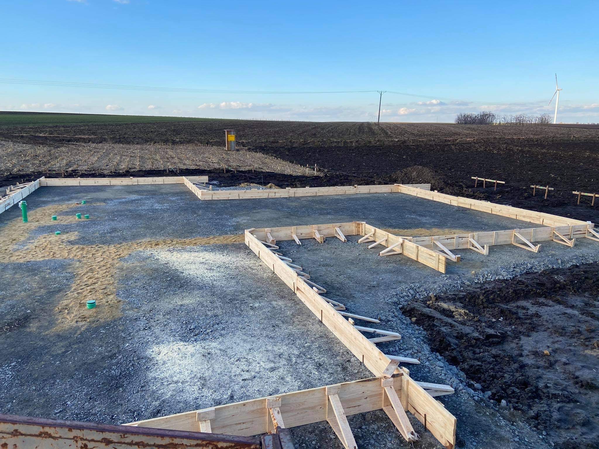 Przygotowany teren pod fundamenty z drewnianym szalunkiem, widoczne zielone rury instalacyjne, w tle pole uprawne i turbina wiatrowa.
