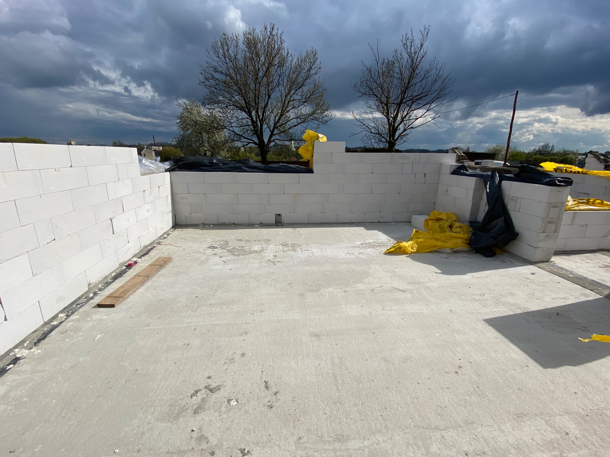 Wnętrze fundamentów domu murowanego z bloczków betonowych, zabezpieczone żółtą folią, widok na pochmurne niebo i drzewa w tle.