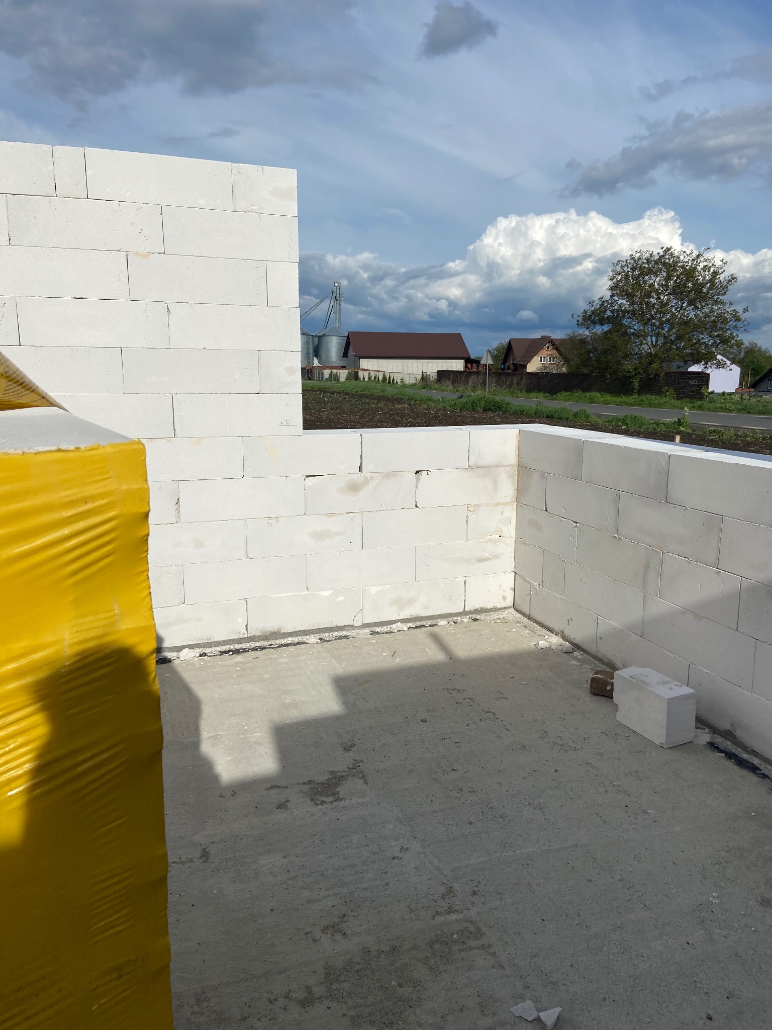 Wznoszone ściany z białych bloczków na fundamencie, widok na wiejskie zabudowania i pochmurne niebo.