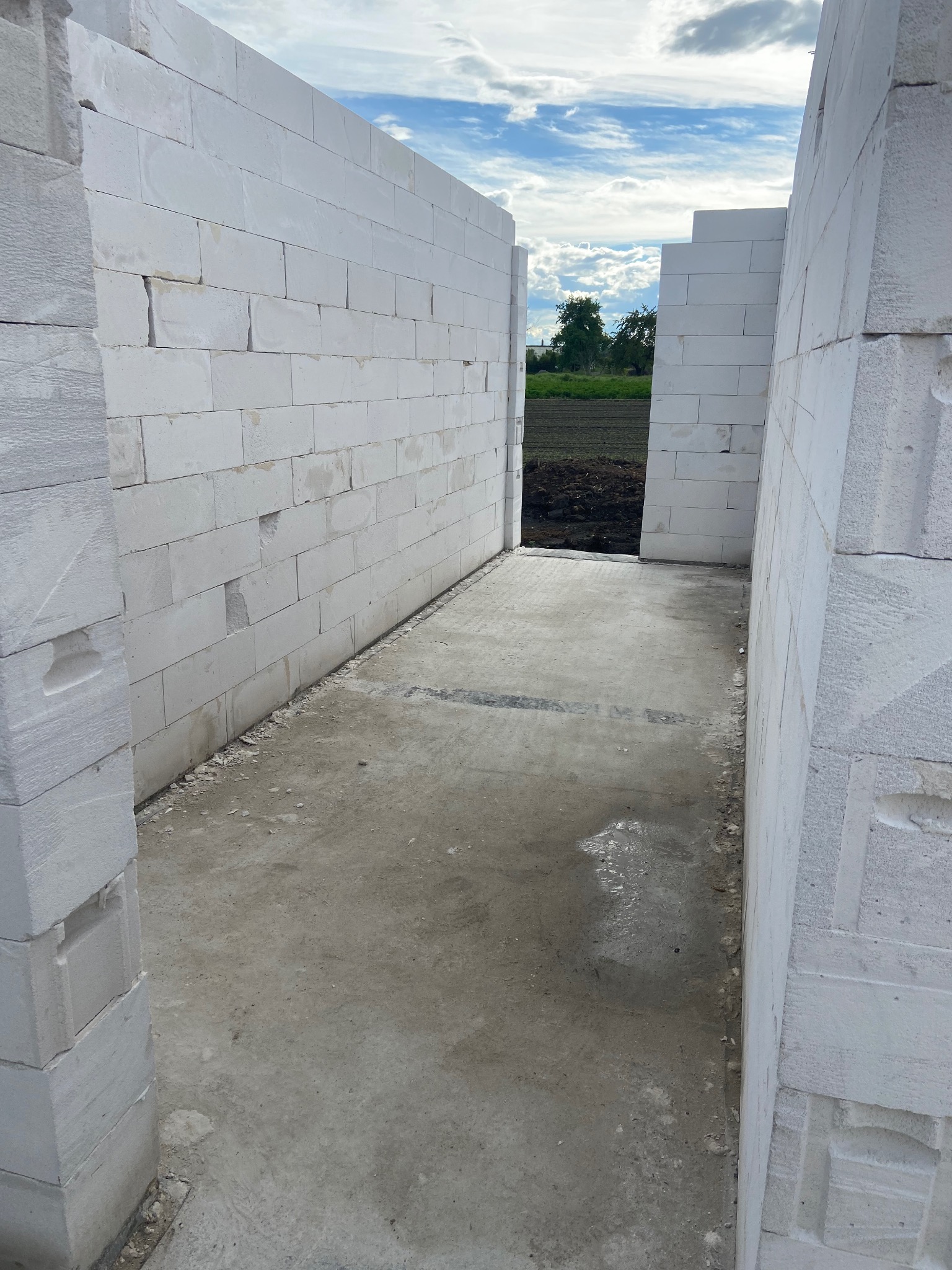 Wnętrze budynku w trakcie budowy, widoczne ściany z białych bloczków betonu komórkowego i betonowa podłoga, na zewnątrz widok na pole i niebo.