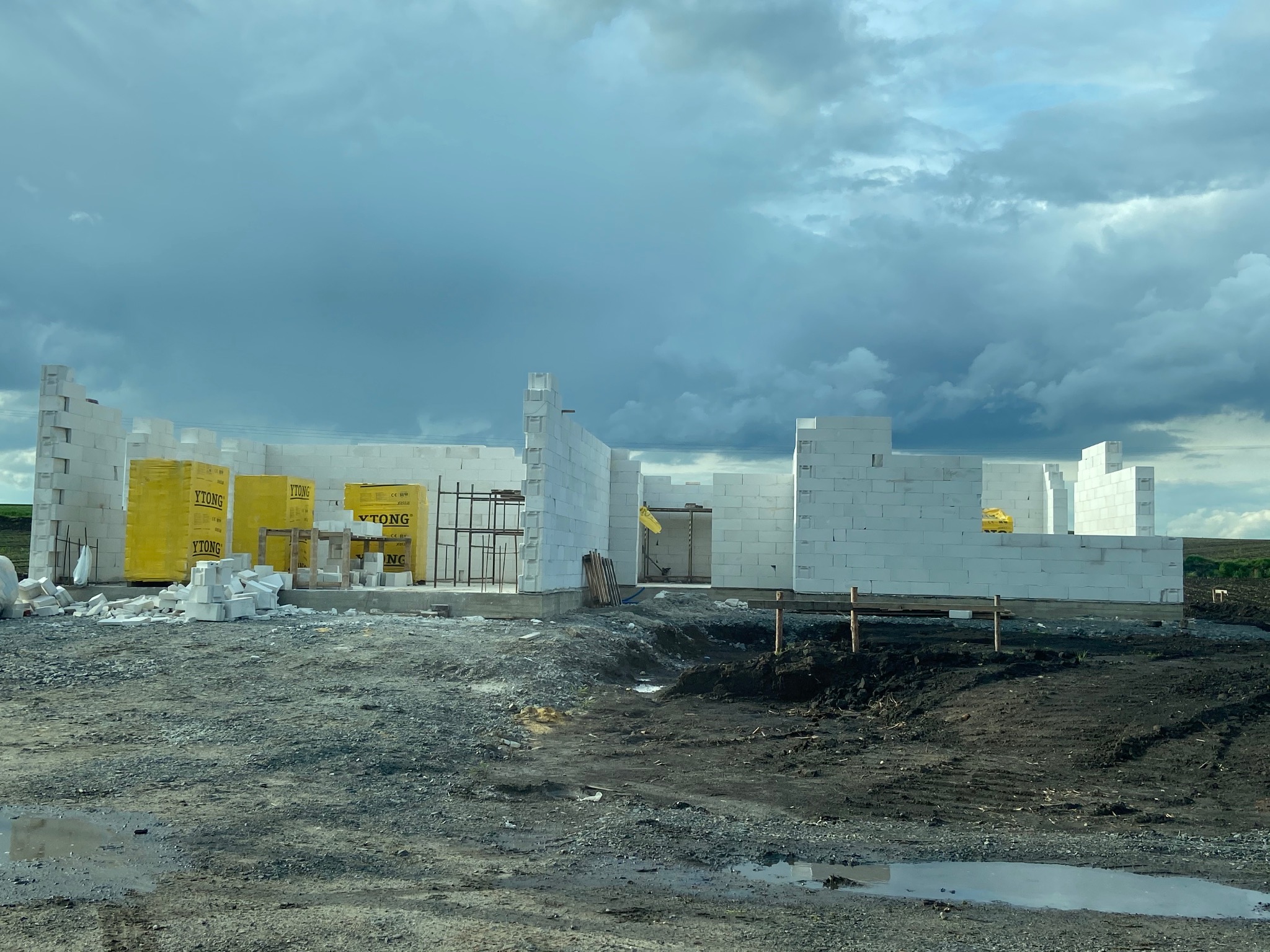 Wznoszone ściany budynku z białych bloczków Ytong na fundamencie, z widocznymi paletami z bloczkami i mokrym, gliniastym terenem wokół, pod pochmurnym niebem.